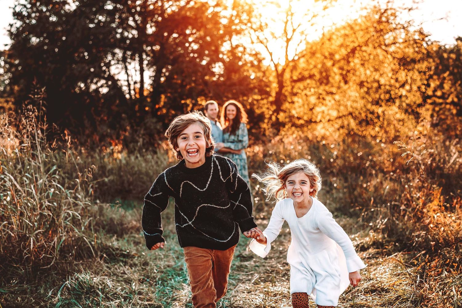 Two joyful children run through a golden autumn field at sunset while their parents smile warmly in the background.