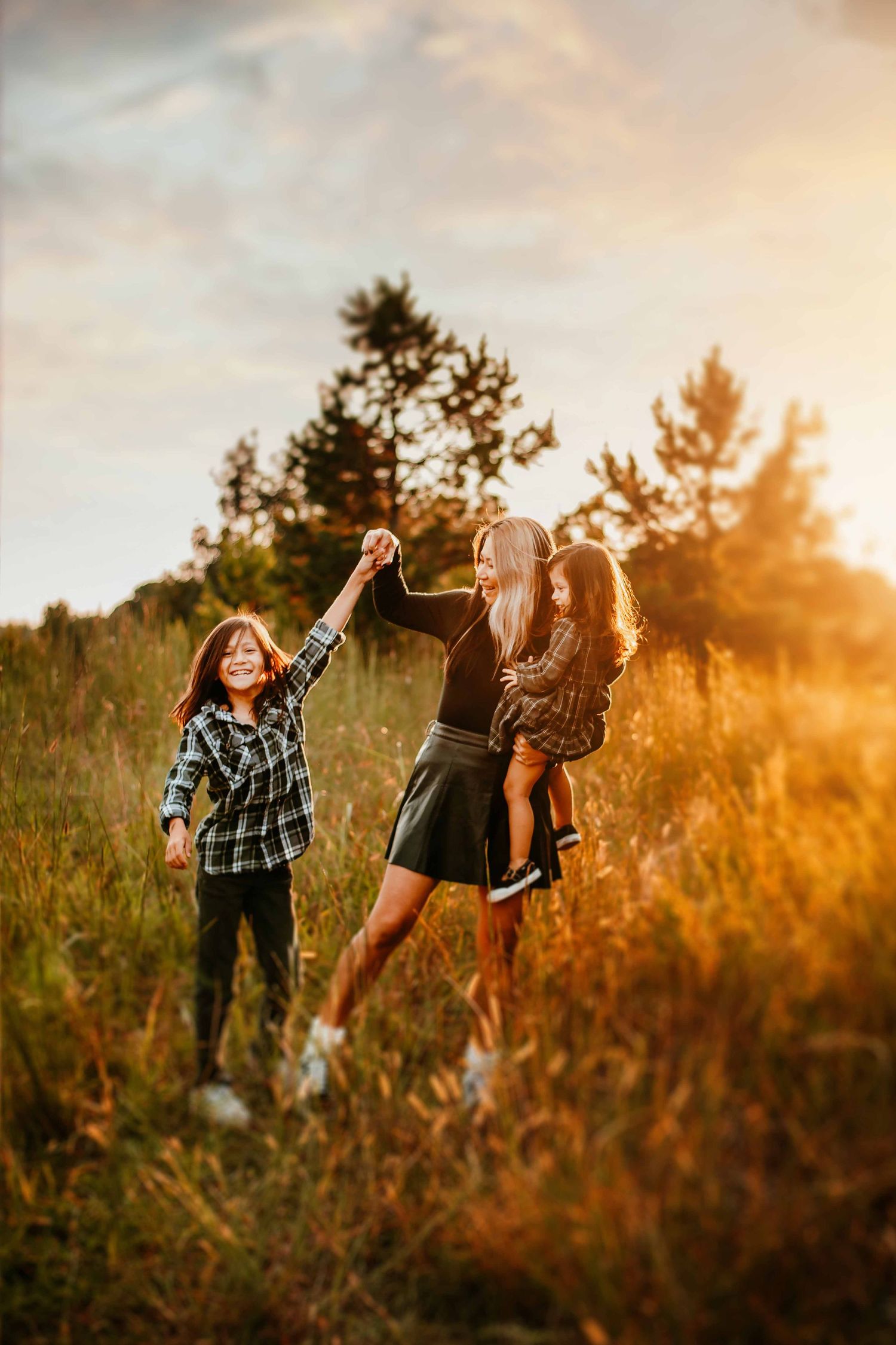 Friends dancing and laughing together in a golden sunlit field at sunset with pine trees in the background.
