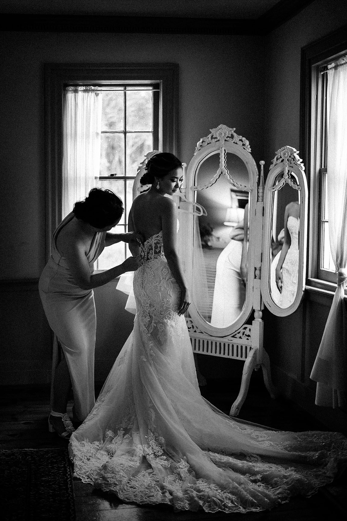 Bride in lace wedding gown being helped by bridesmaid near ornate white mirror in black and white photo.