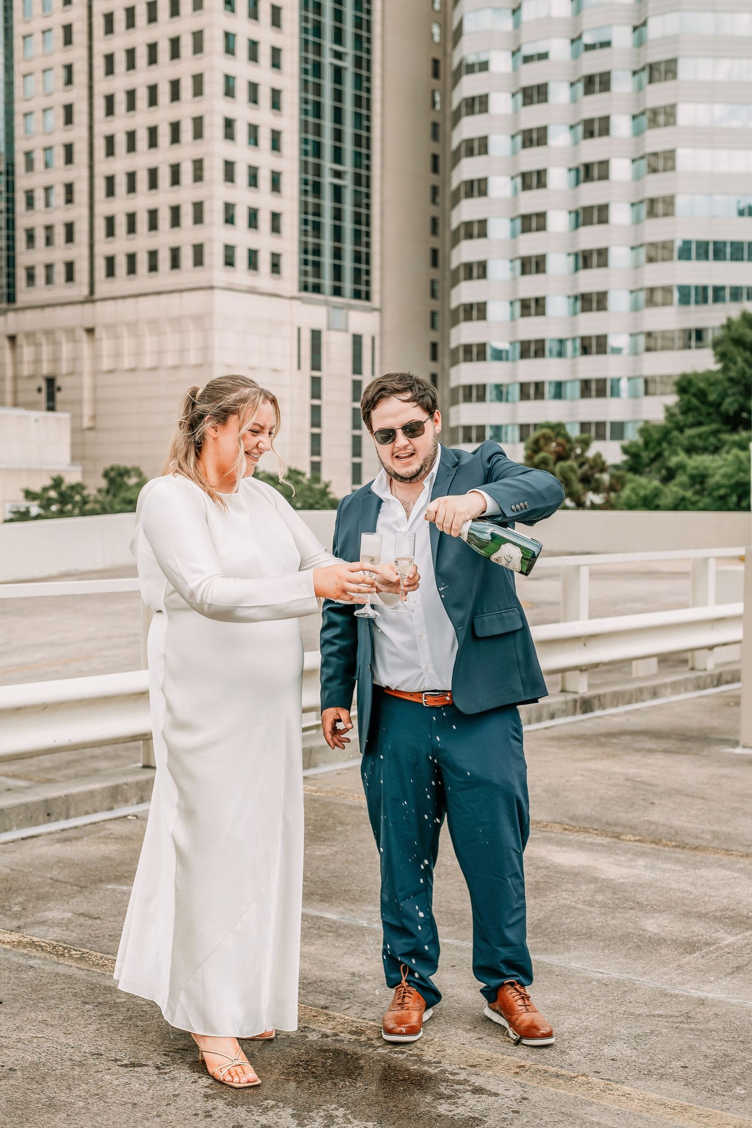 Bride and groom celebrate on urban rooftop, popping champagne in city skyline setting.