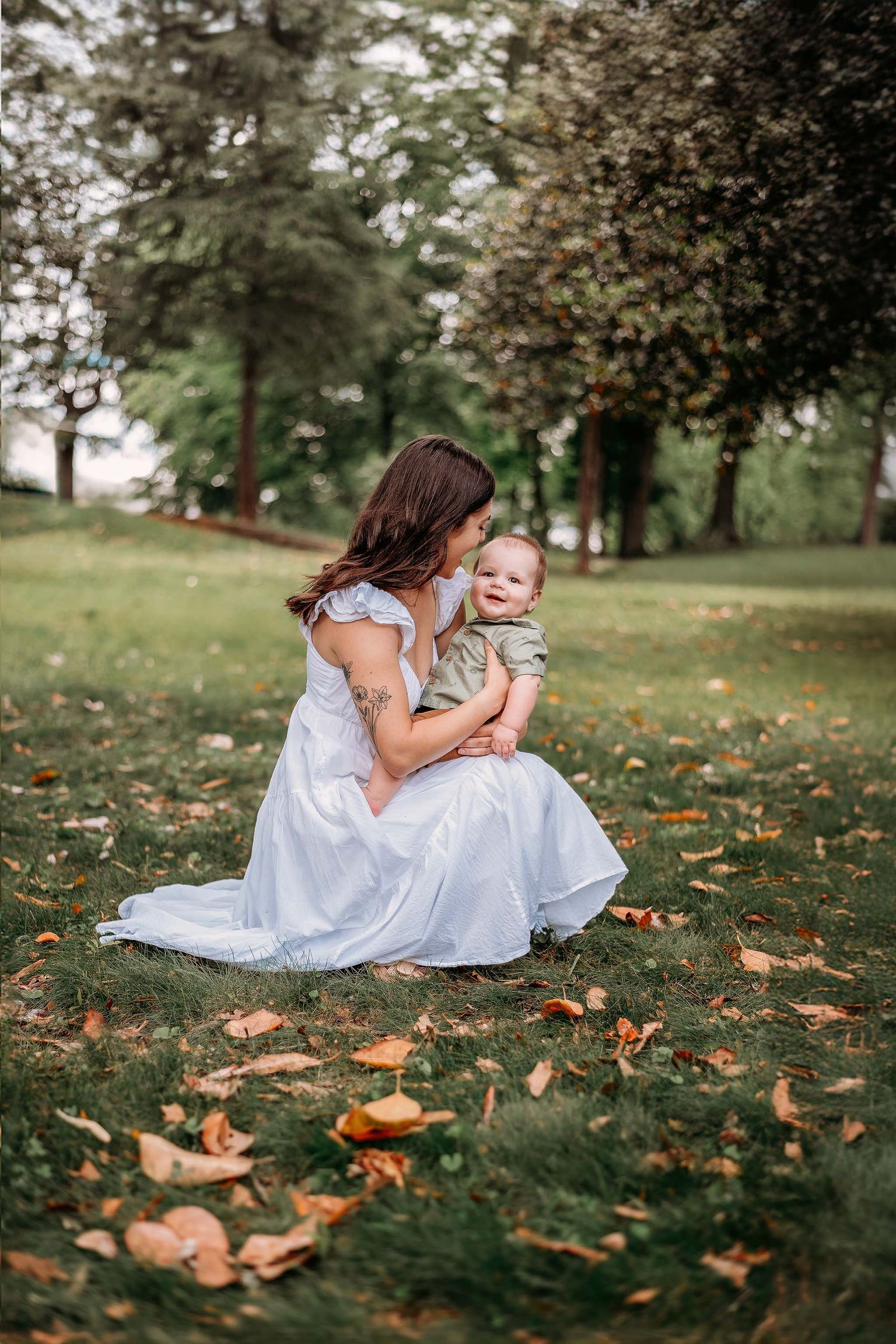 A mother in a white dress sits on green grass holding her baby in a lush park with autumn leaves.