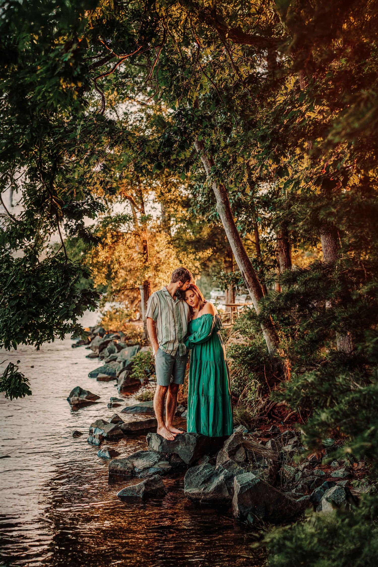 Couple embracing on rocky shoreline surrounded by lush green trees during golden hour sunset.