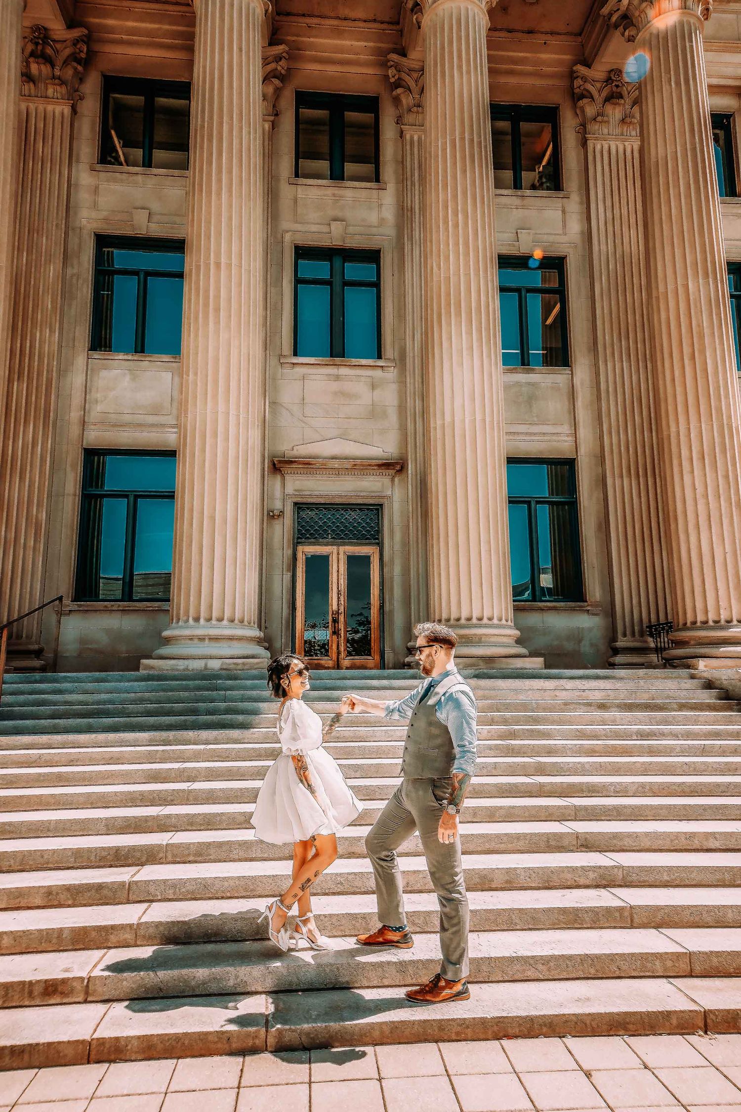 Couple holding hands on steps of a grand neoclassical building with tall columns, woman in white dress, man in gray suit.
