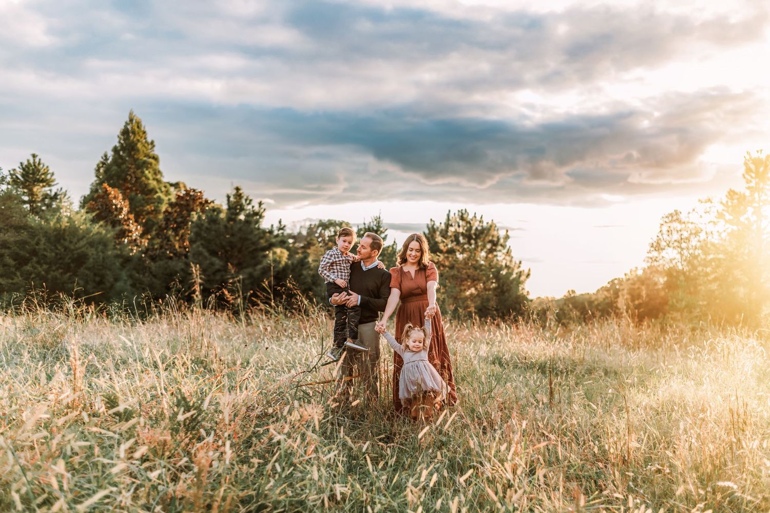 Family standing in golden sunlit field during sunset with trees in background.