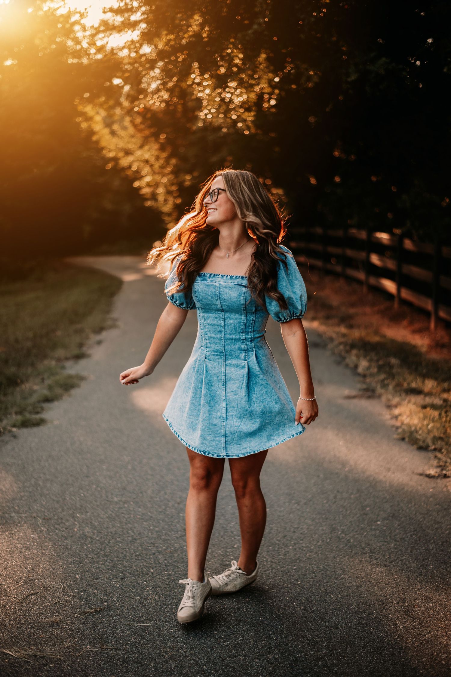 Woman in blue denim dress twirling on path during golden hour.