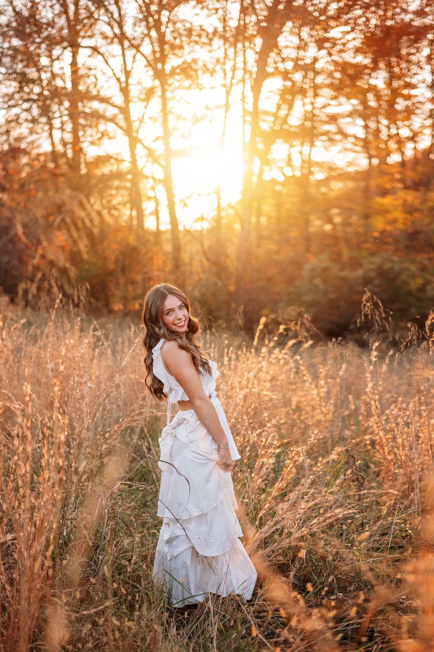 A white dress flows in a golden field at sunset as warm light filters through autumn trees in the background.