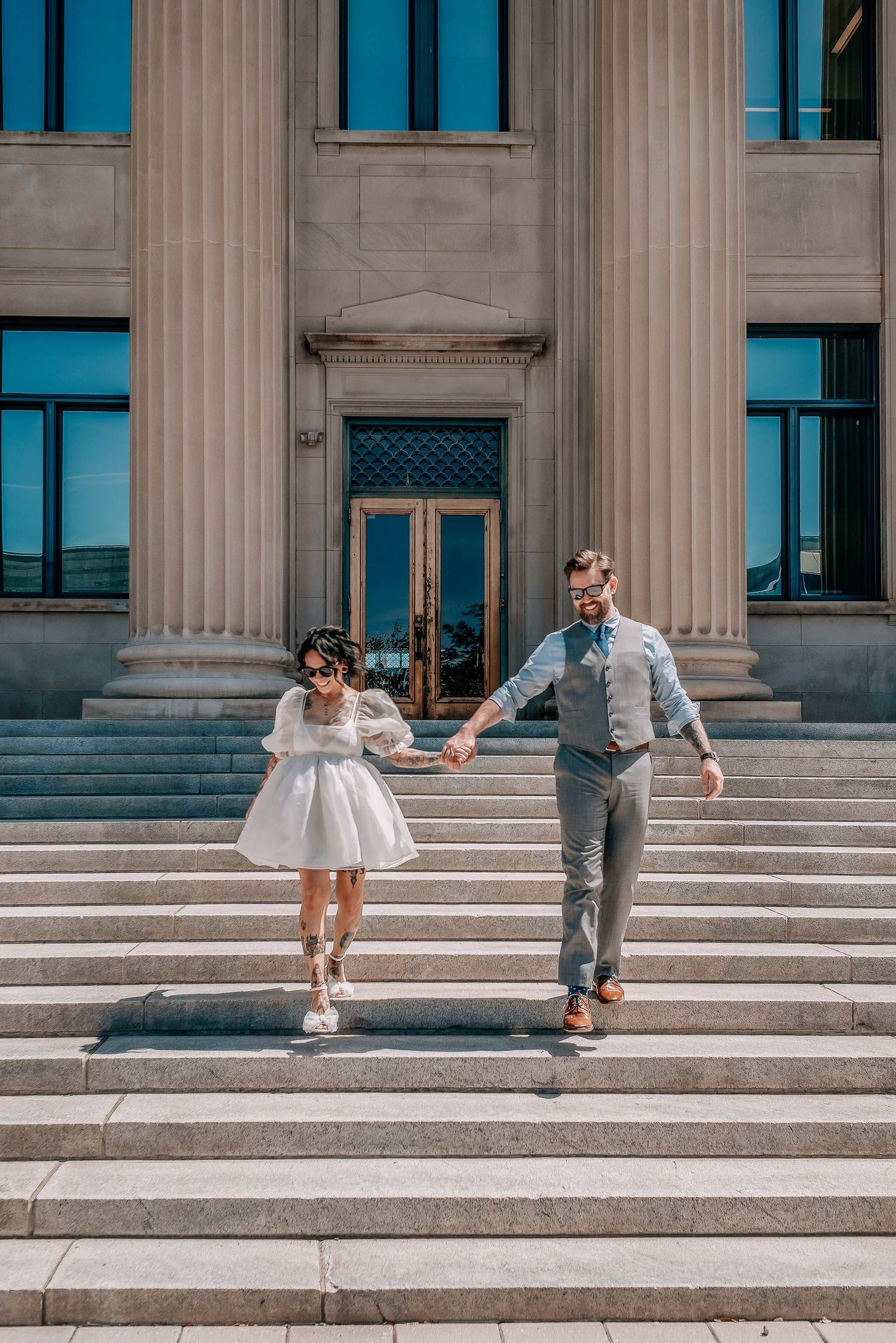 Bride in short white dress and groom in gray suit hold hands walking down stone steps of elegant building.