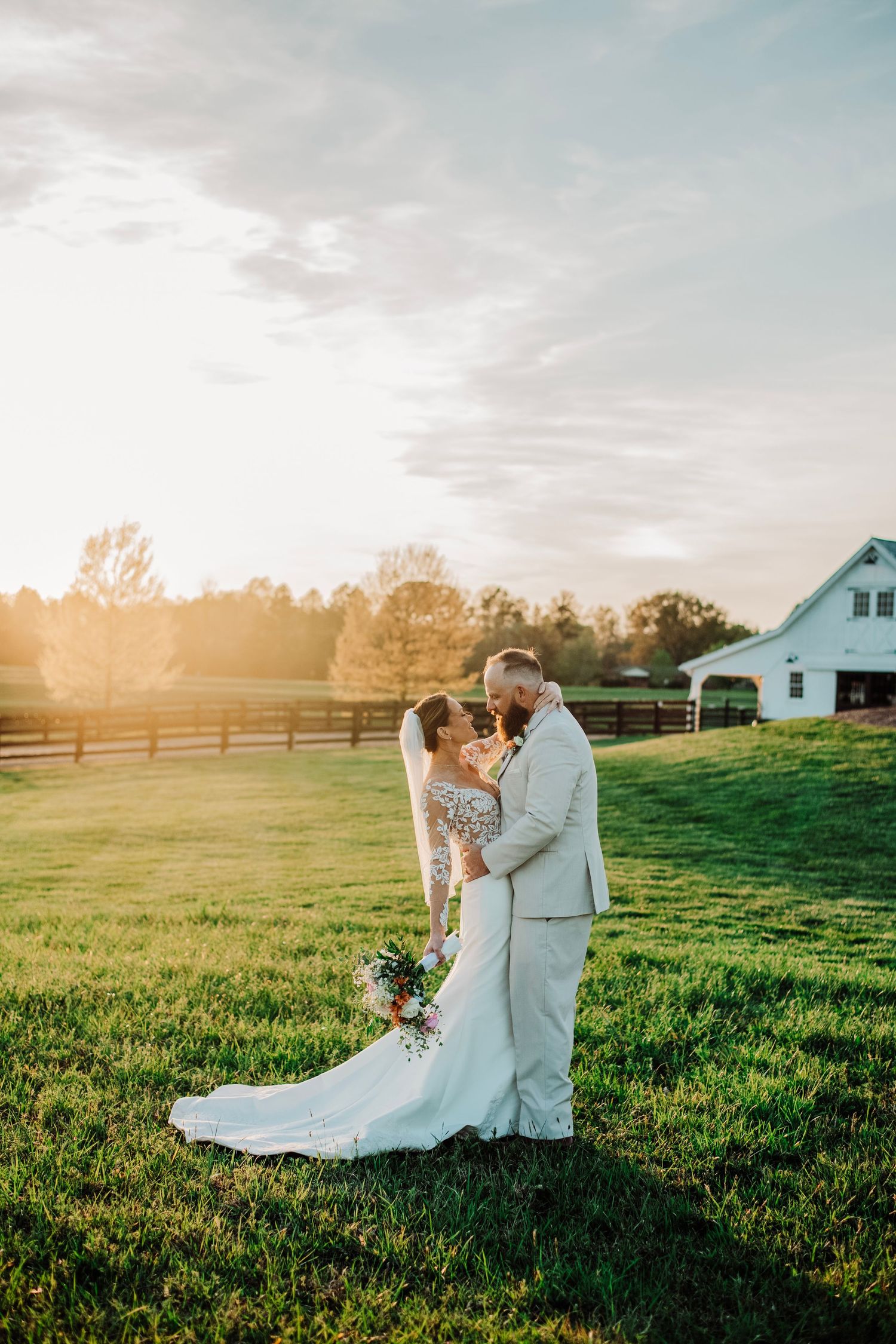 Bride and groom share romantic sunset kiss in green field with white barn and wooden fence in background.
