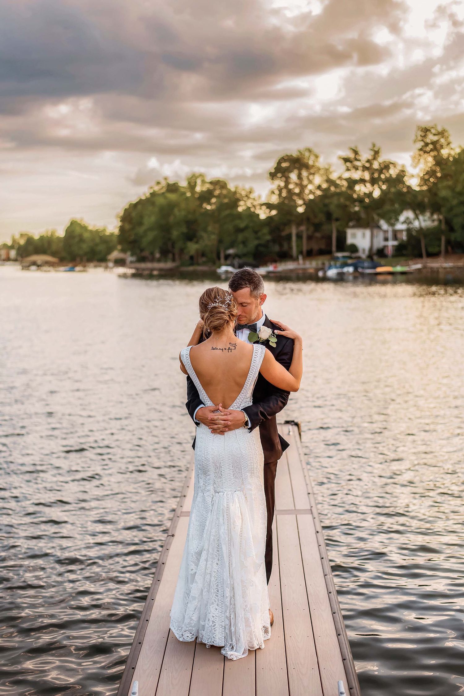 Bride and groom embrace on a lakeside dock at sunset, bride in lace gown with back tattoo, groom in dark suit.