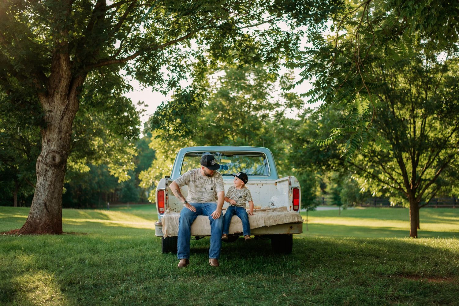 Two people sitting on the tailgate of a vintage white pickup truck under lush green trees in a sunny park.