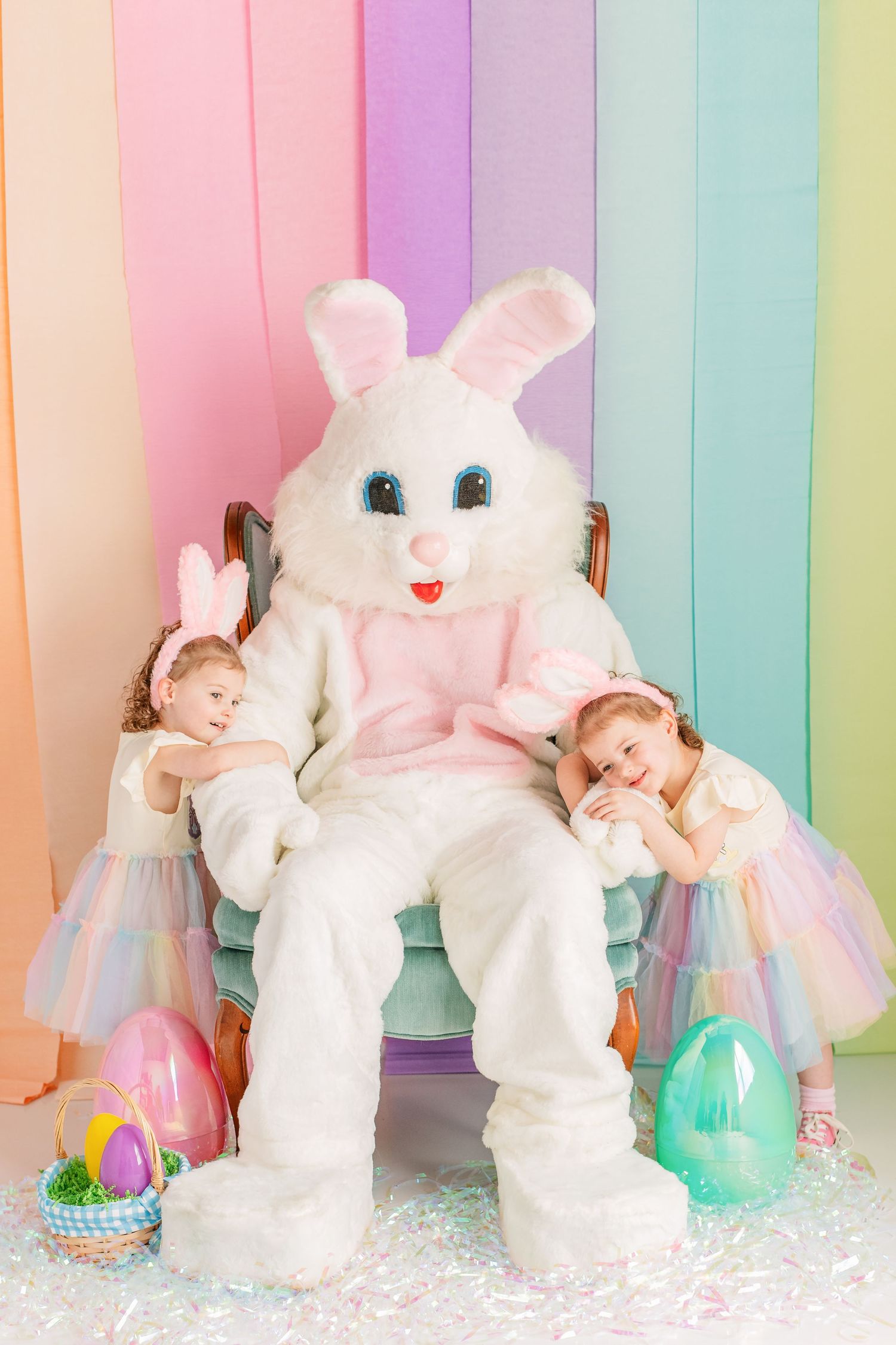 Adorable white bunny plush toys arranged against a pastel rainbow striped background with Easter eggs and glitter.