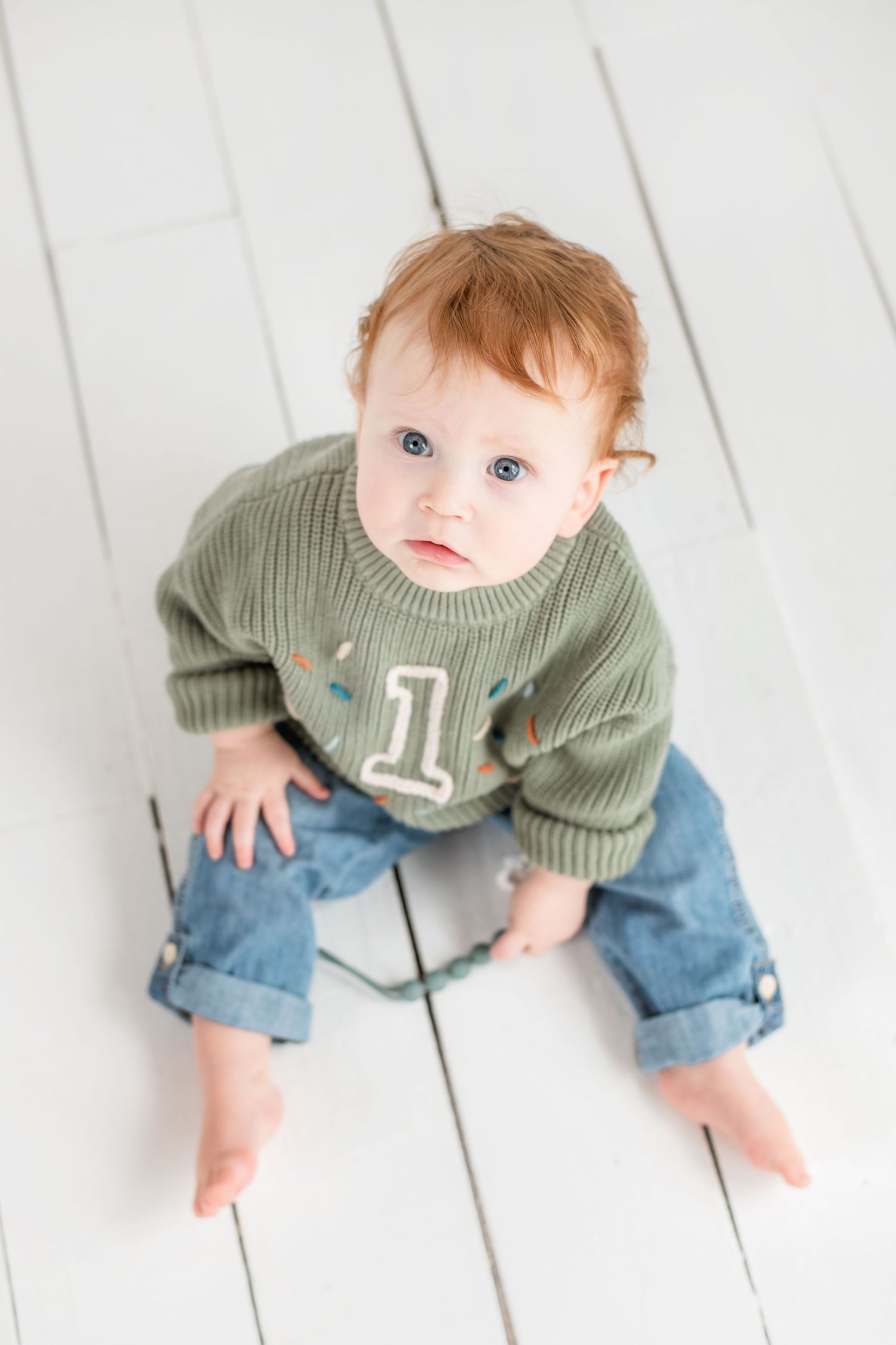 Baby in sage green sweater with number 1 and rolled-up jeans sits on white wooden floor.
