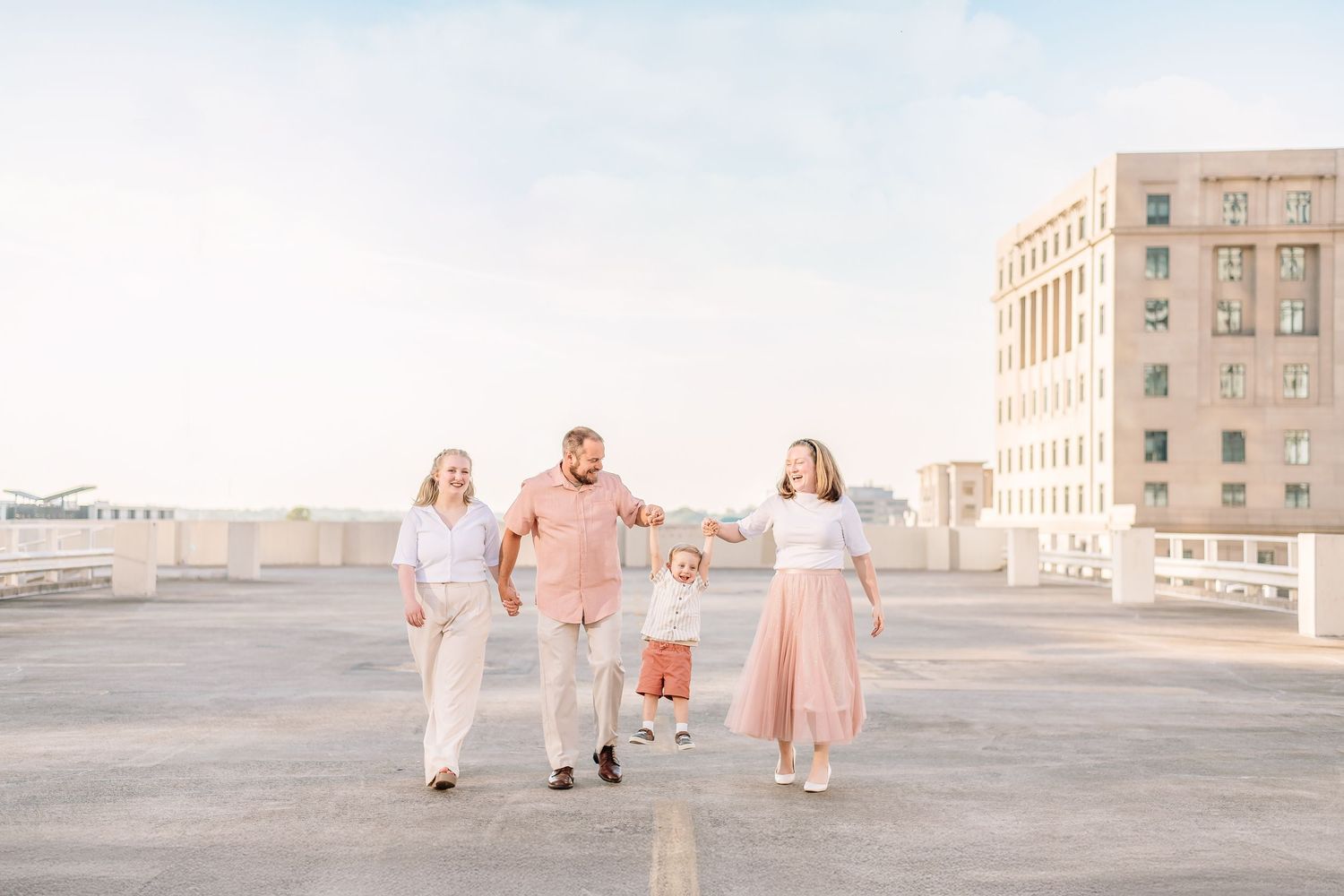 A family wearing coordinated pink and white outfits poses on an urban rooftop at sunset.