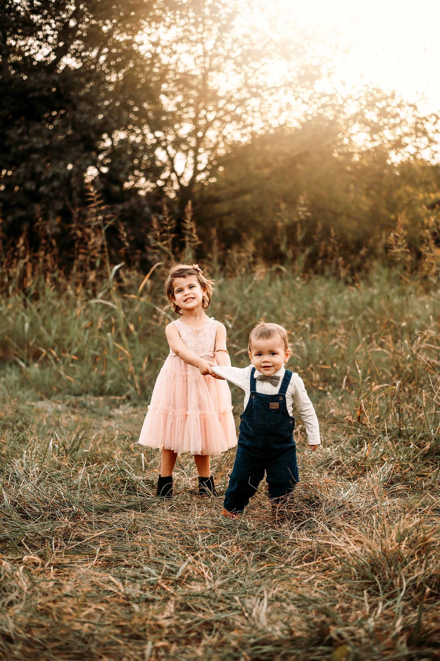 Two young children holding hands in a sunlit field, girl in pink dress and boy in navy overalls with bow tie.