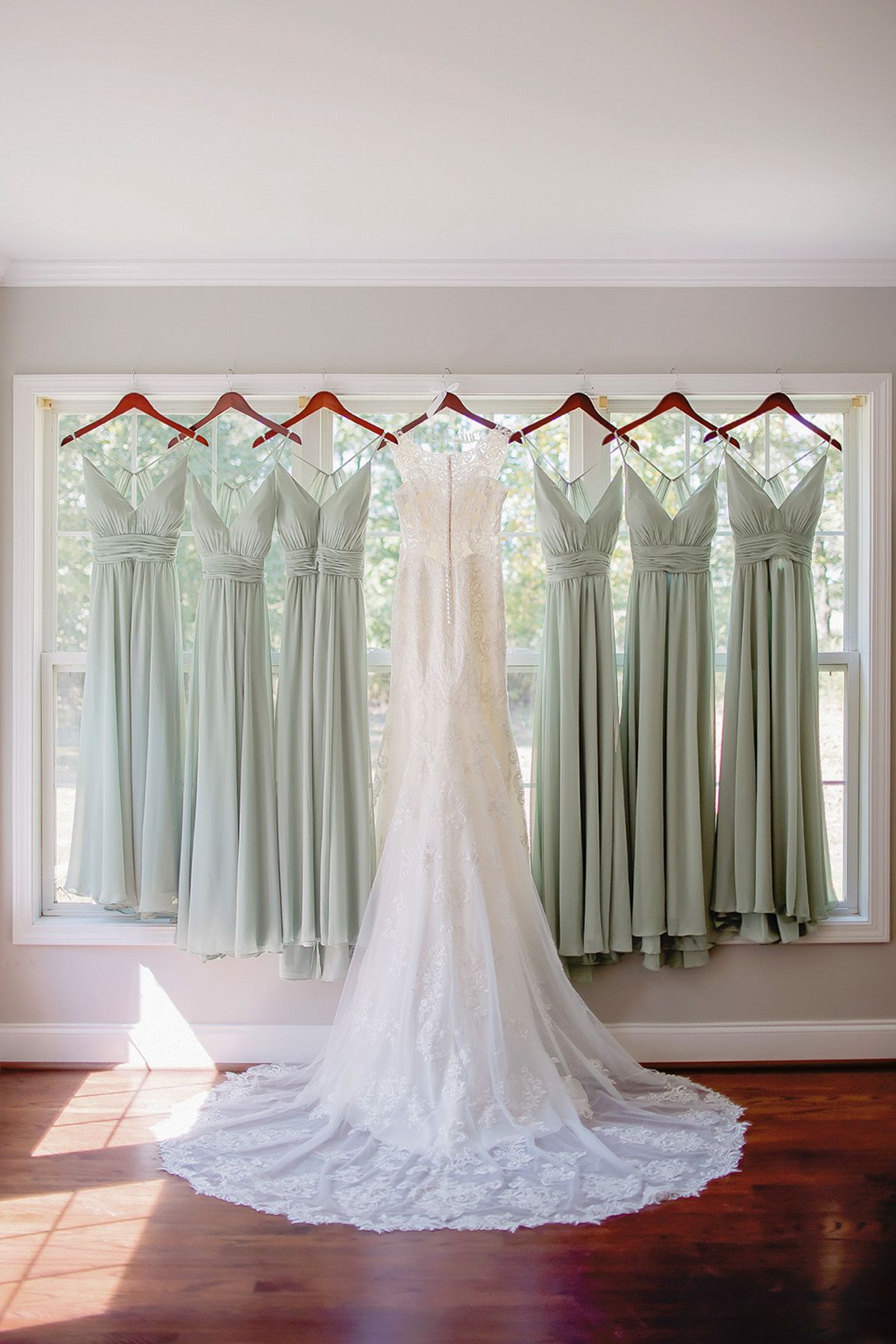 White wedding gown with lace train displayed beside sage green bridesmaid dresses hanging on wooden hangers in a sunlit window.