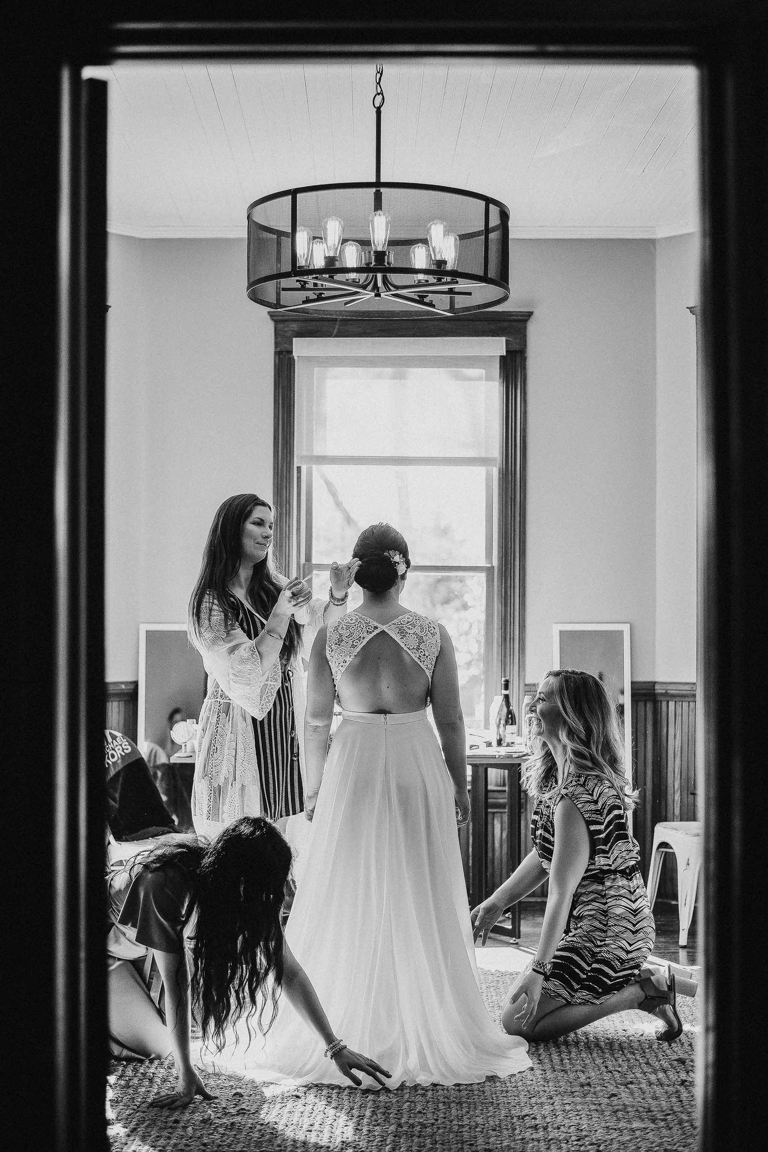 Black and white photo of bride being helped into wedding dress by bridesmaids in elegant room with chandelier.
