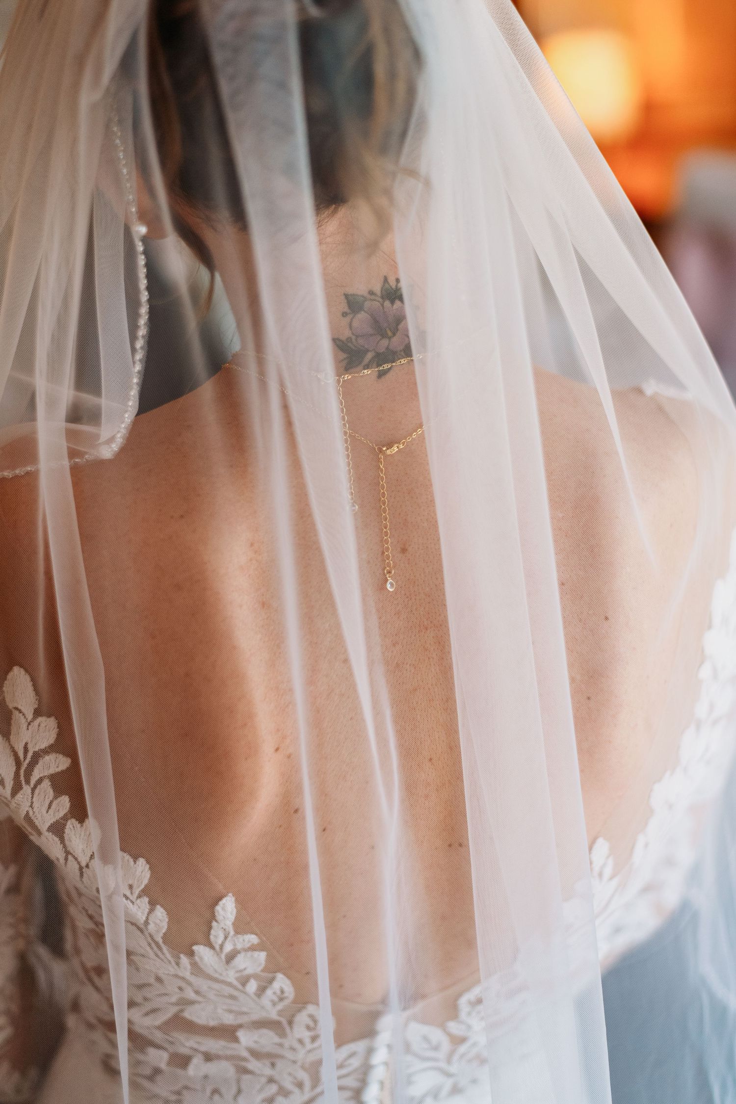 Close-up of bride in lace wedding dress with veil, showing gold back necklace and floral tattoo on shoulder.