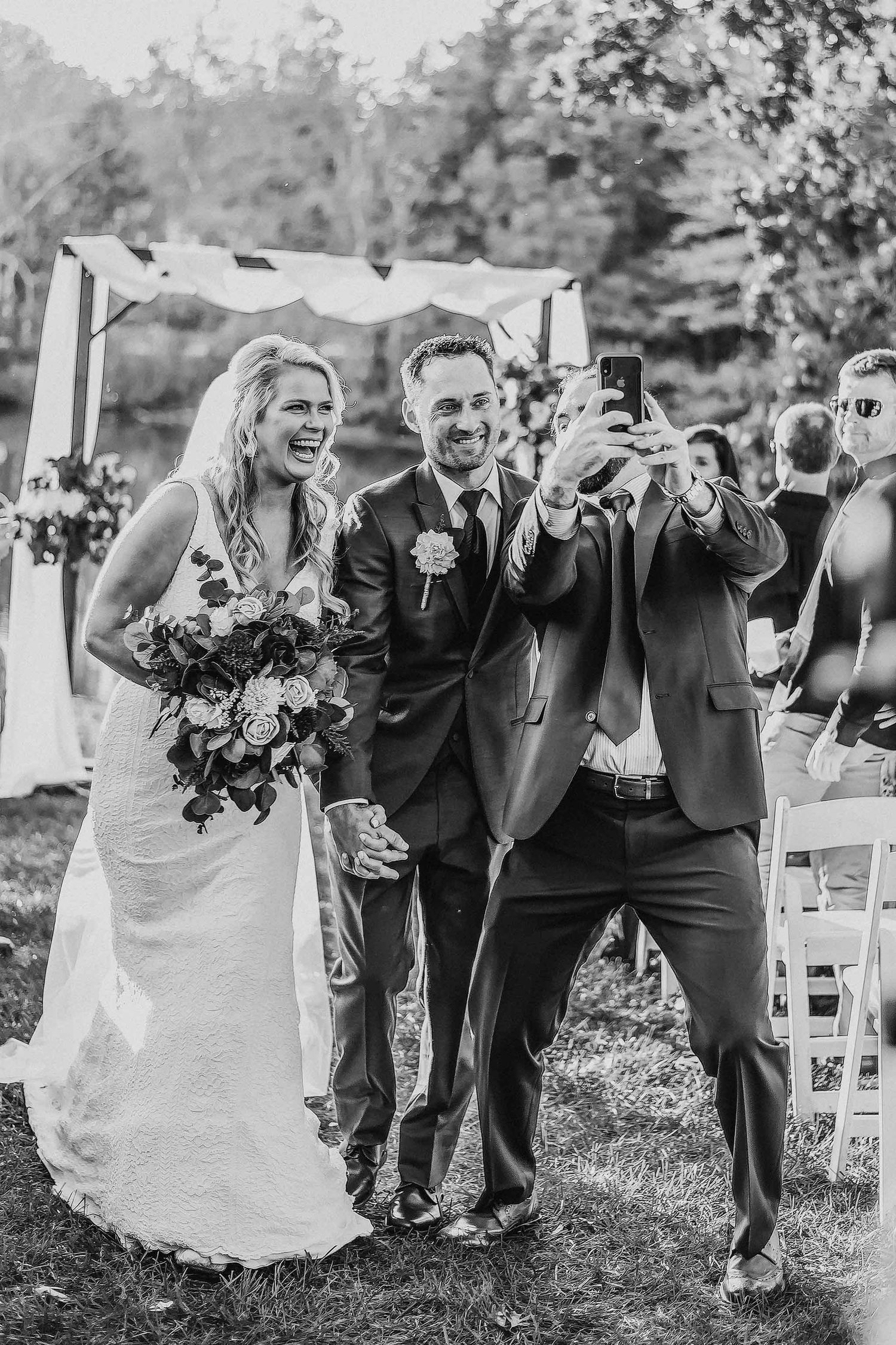 Black and white photo of a joyful bride and groom taking a selfie after their outdoor wedding ceremony.
