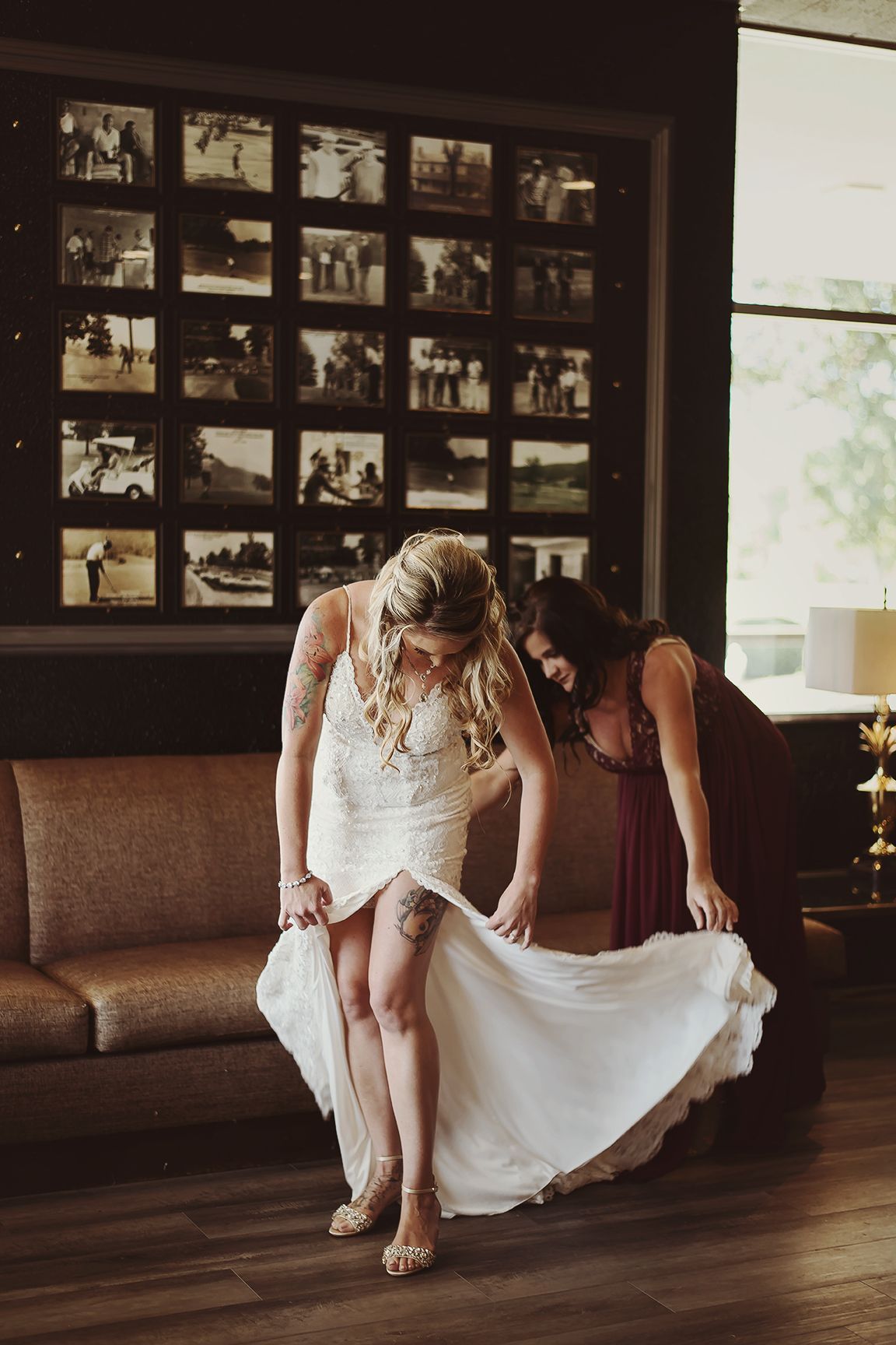 A bride in a white lace wedding dress adjusts her gown while sitting on a brown couch with framed photos on the wall.