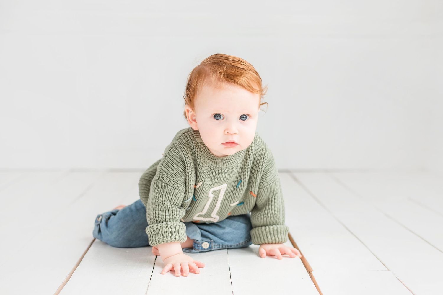Toddler wearing sage green sweater and blue jeans sits on white wooden floor.