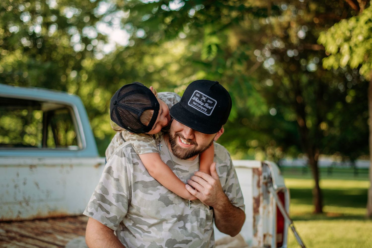 A smiling man in a camo shirt and black cap carries a child on his back outdoors near a vintage white truck.