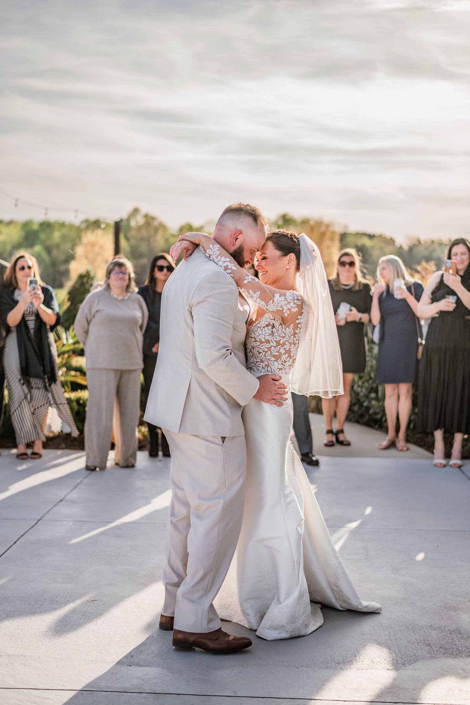 Bride and groom share first dance kiss outdoors at wedding reception with guests applauding in background.