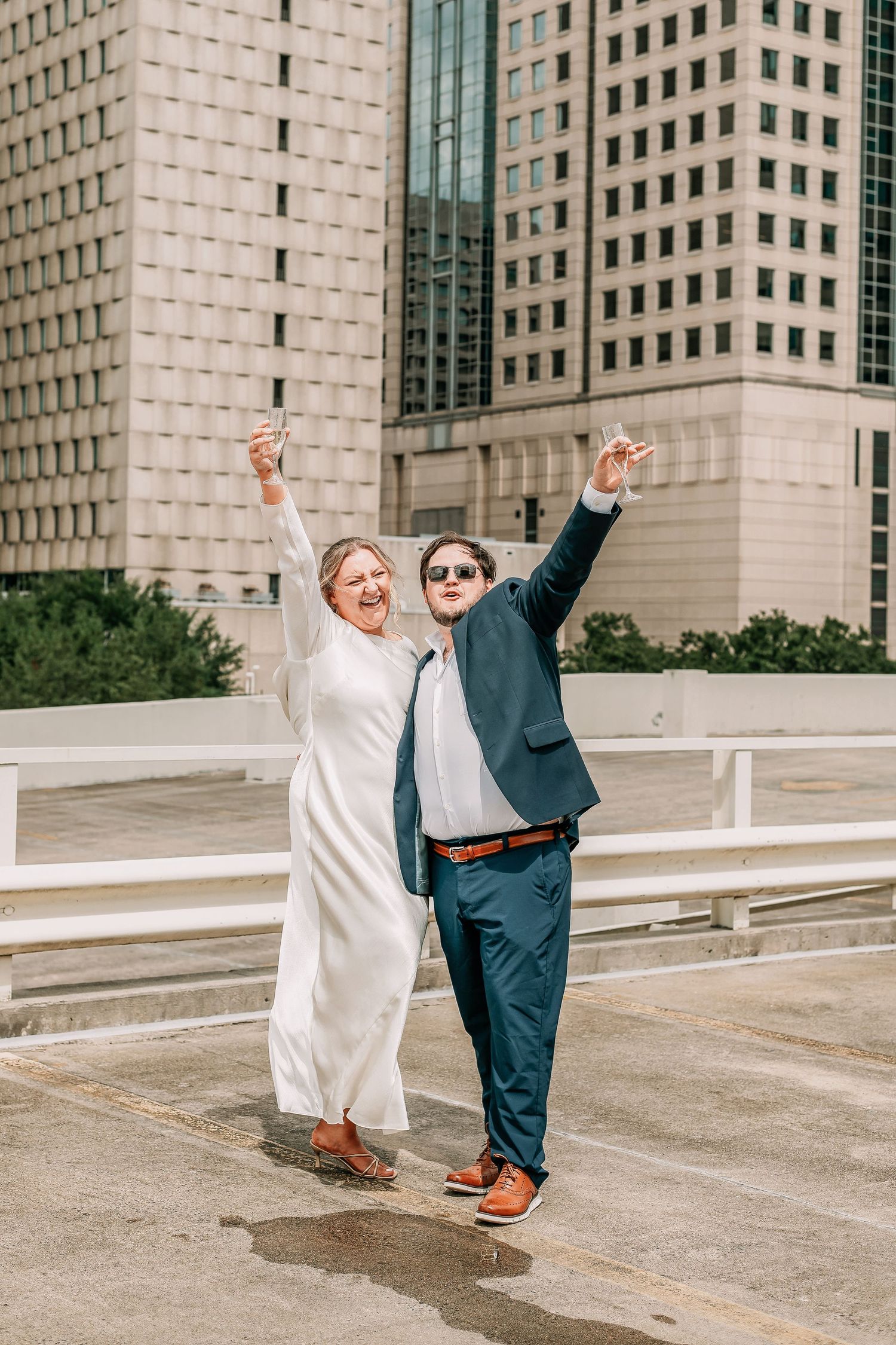 Joyful newlyweds celebrate on a rooftop parking garage, arms raised, wearing white dress and navy suit with city buildings behind.