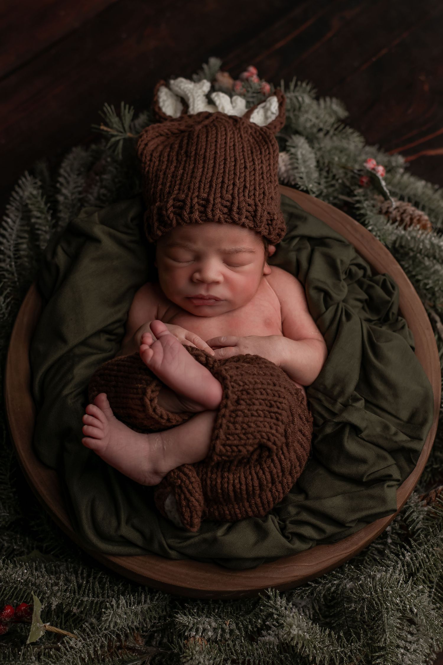 Sleeping newborn in brown knit hat and pants nestled in green fabric with evergreen branches and floral accents.
