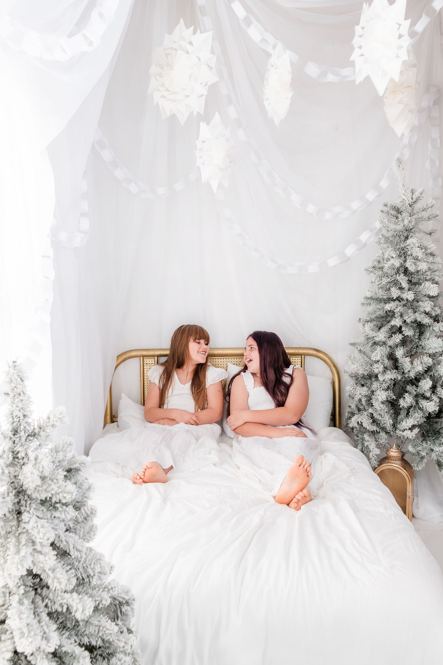 Two people in white dresses lying on a bed surrounded by snowy white Christmas trees in a dreamy winter setting.