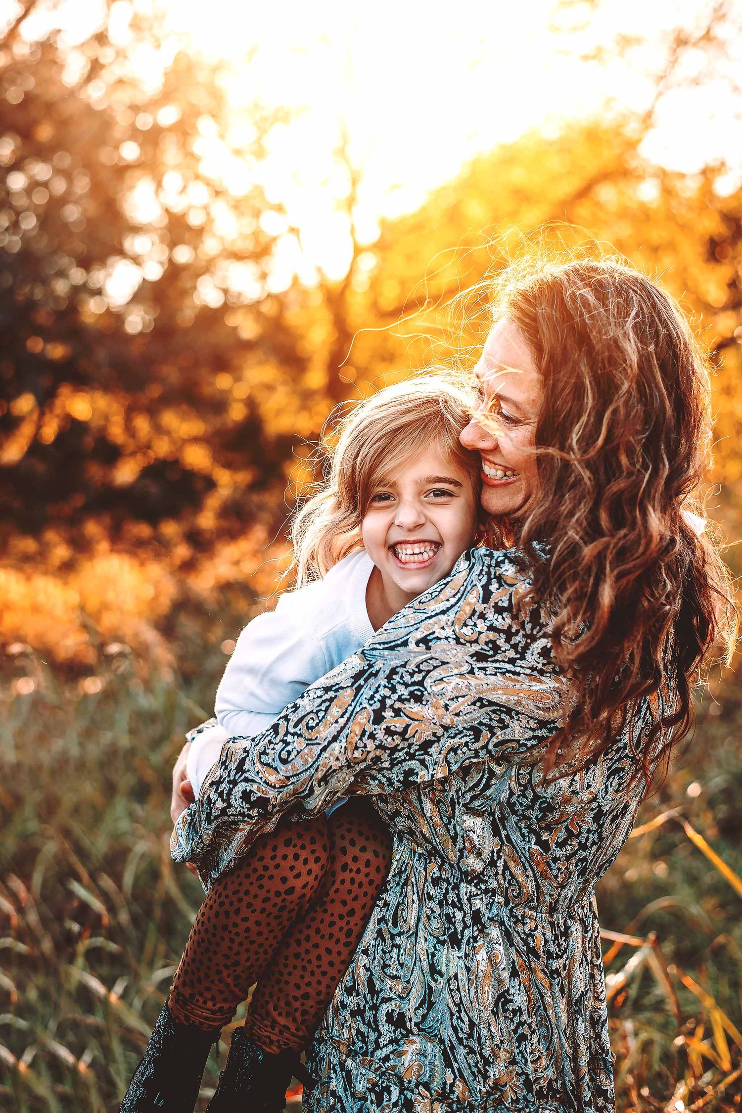 A joyful mother hugging her laughing daughter outdoors in golden sunset light among autumn foliage.
