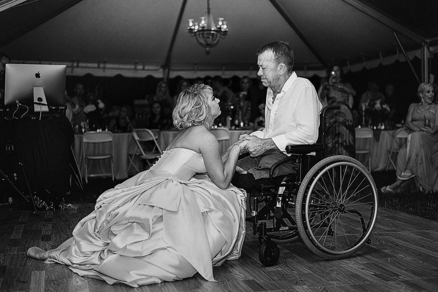 Bride kneels beside groom in wheelchair during emotional first dance at outdoor wedding reception under tent.