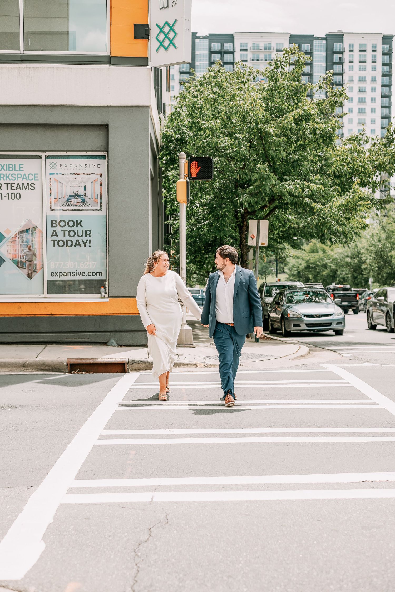 Couple in formal attire crossing a city street crosswalk, with urban buildings and green trees in the background.