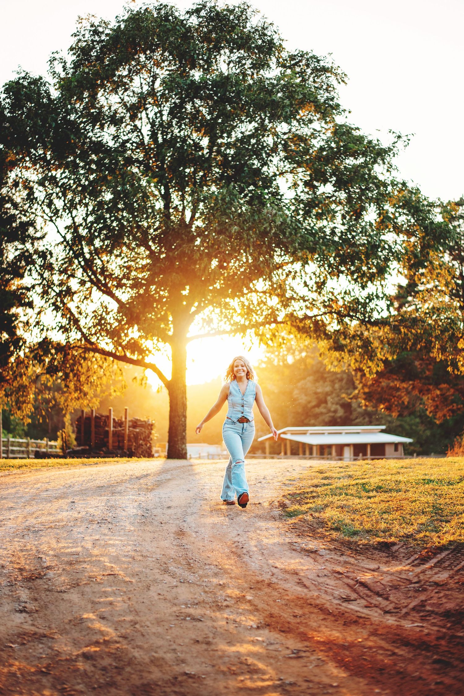 A child in a blue dress walks down a sunlit dirt path beneath trees during golden hour at sunset.