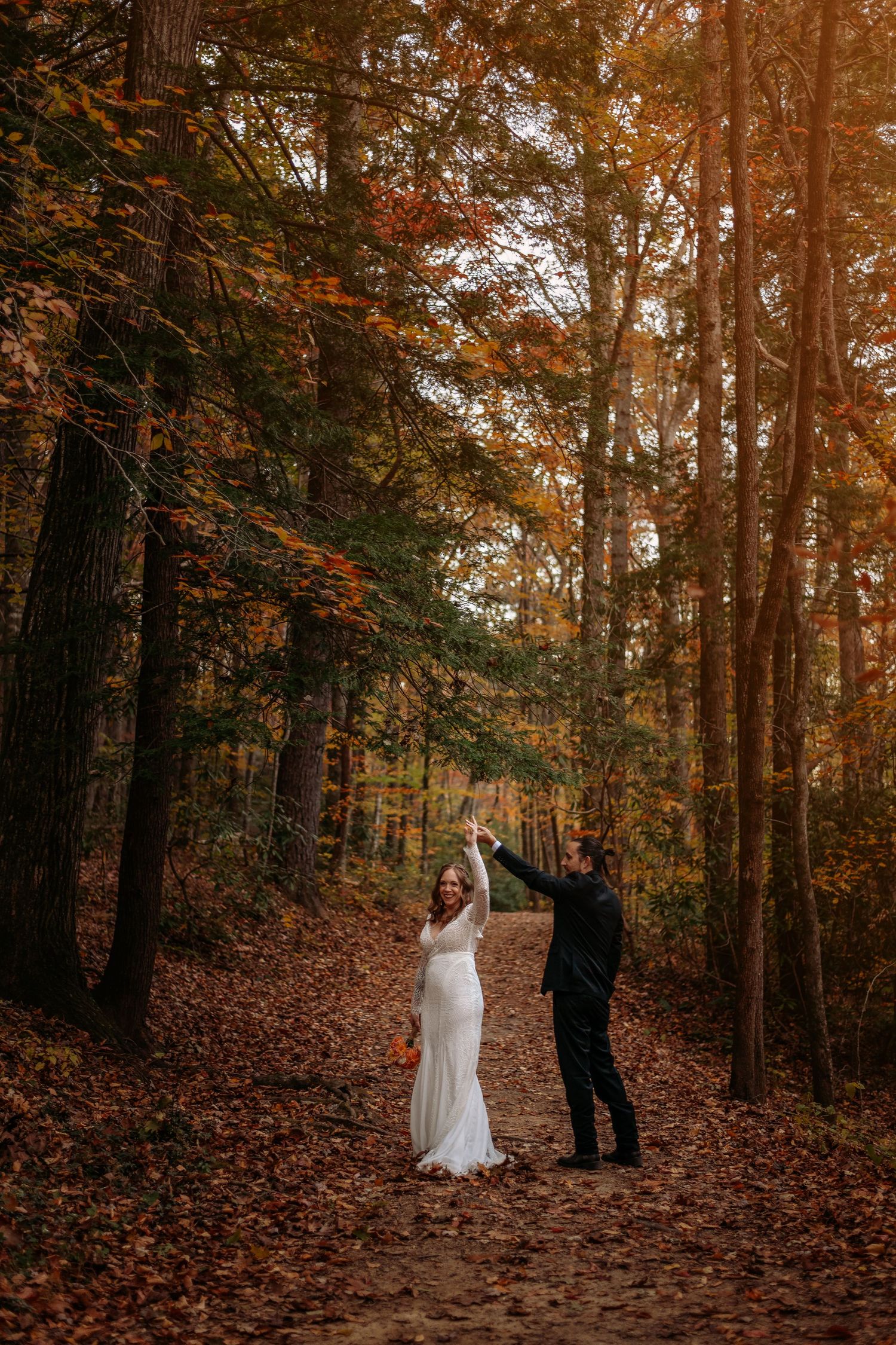 Bride and groom dancing on a forest path surrounded by vibrant autumn foliage in a romantic fall wedding photo.