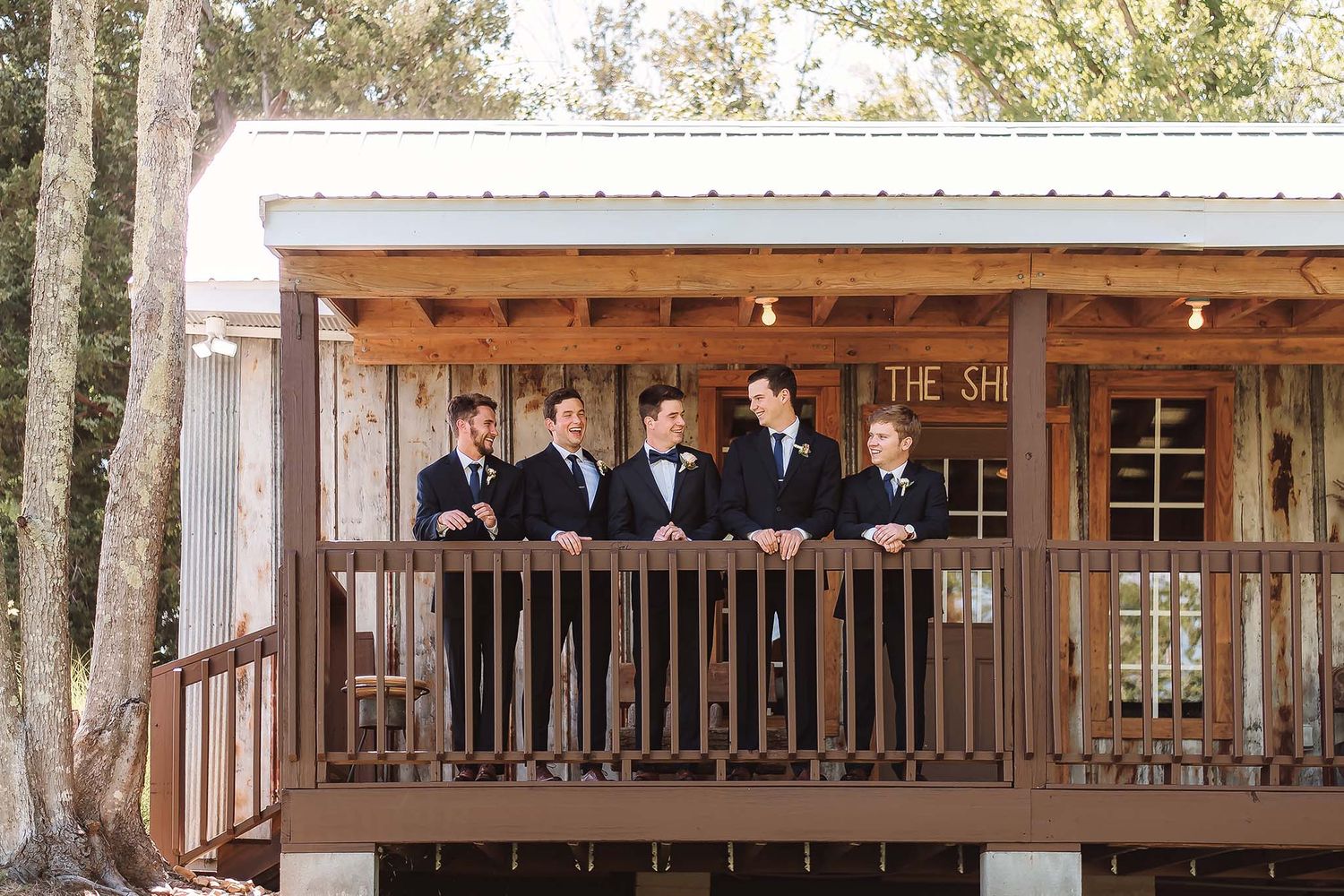 Groomsmen in navy suits stand on a rustic wooden deck at an outdoor wedding venue surrounded by trees.