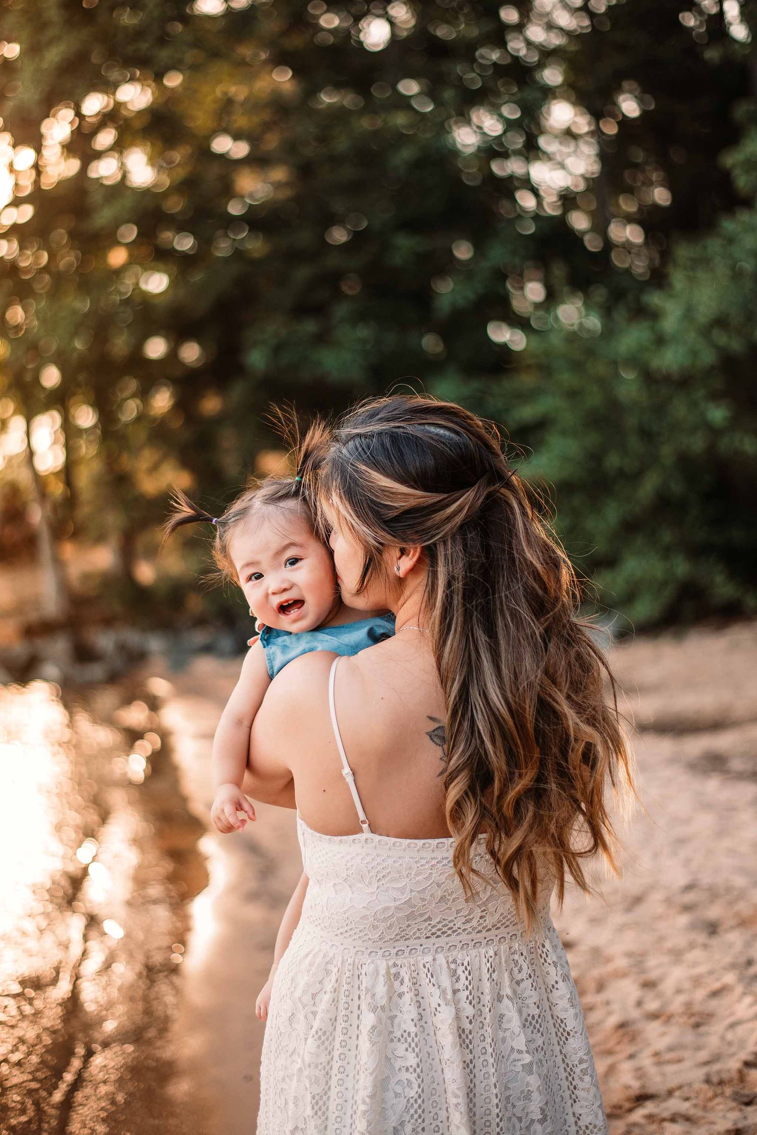 A mother in a white lace dress holds her smiling baby outdoors in golden hour light near a forest path.