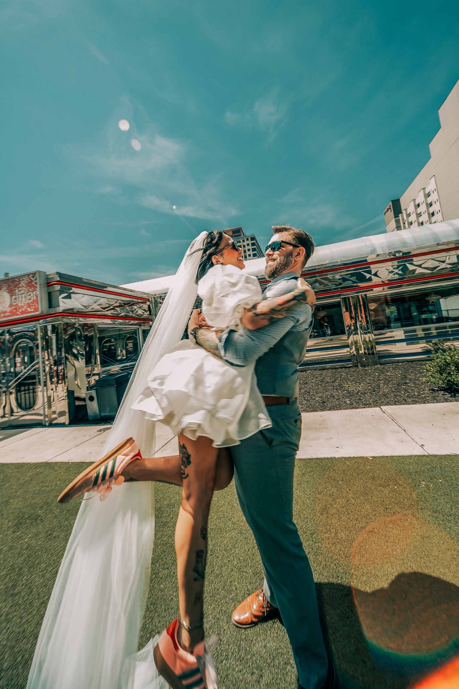 A groom in a blue suit lifts his bride in a short white dress and veil outdoors on a sunny day.