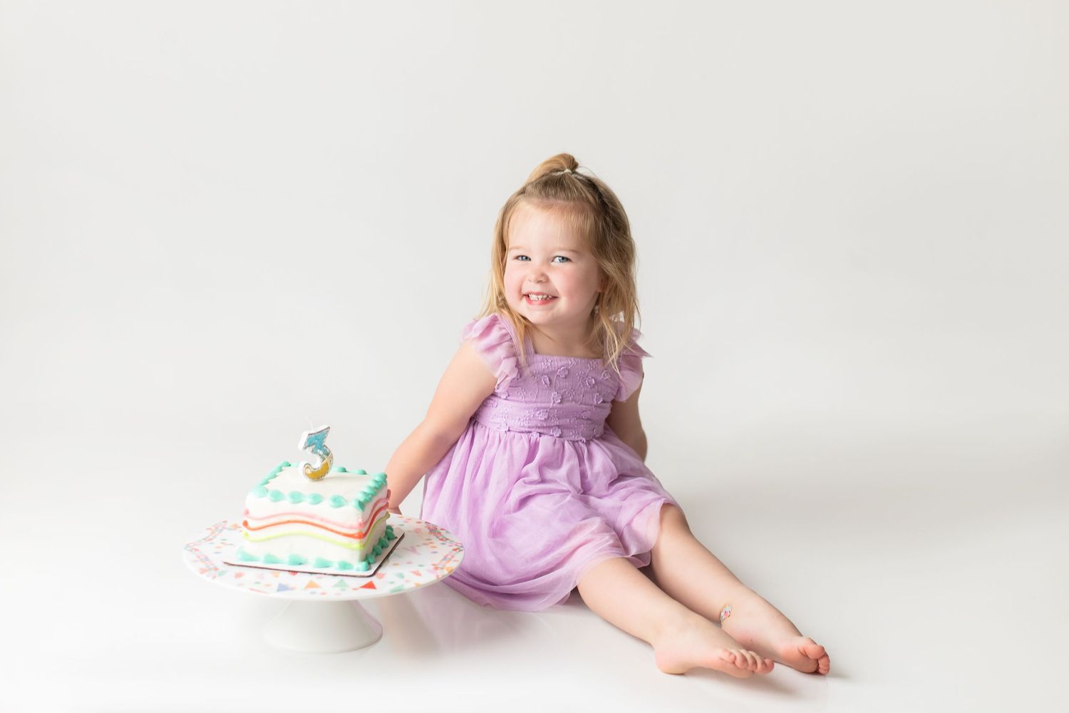 Young child in purple dress sits on white backdrop with decorative pillow beside them.