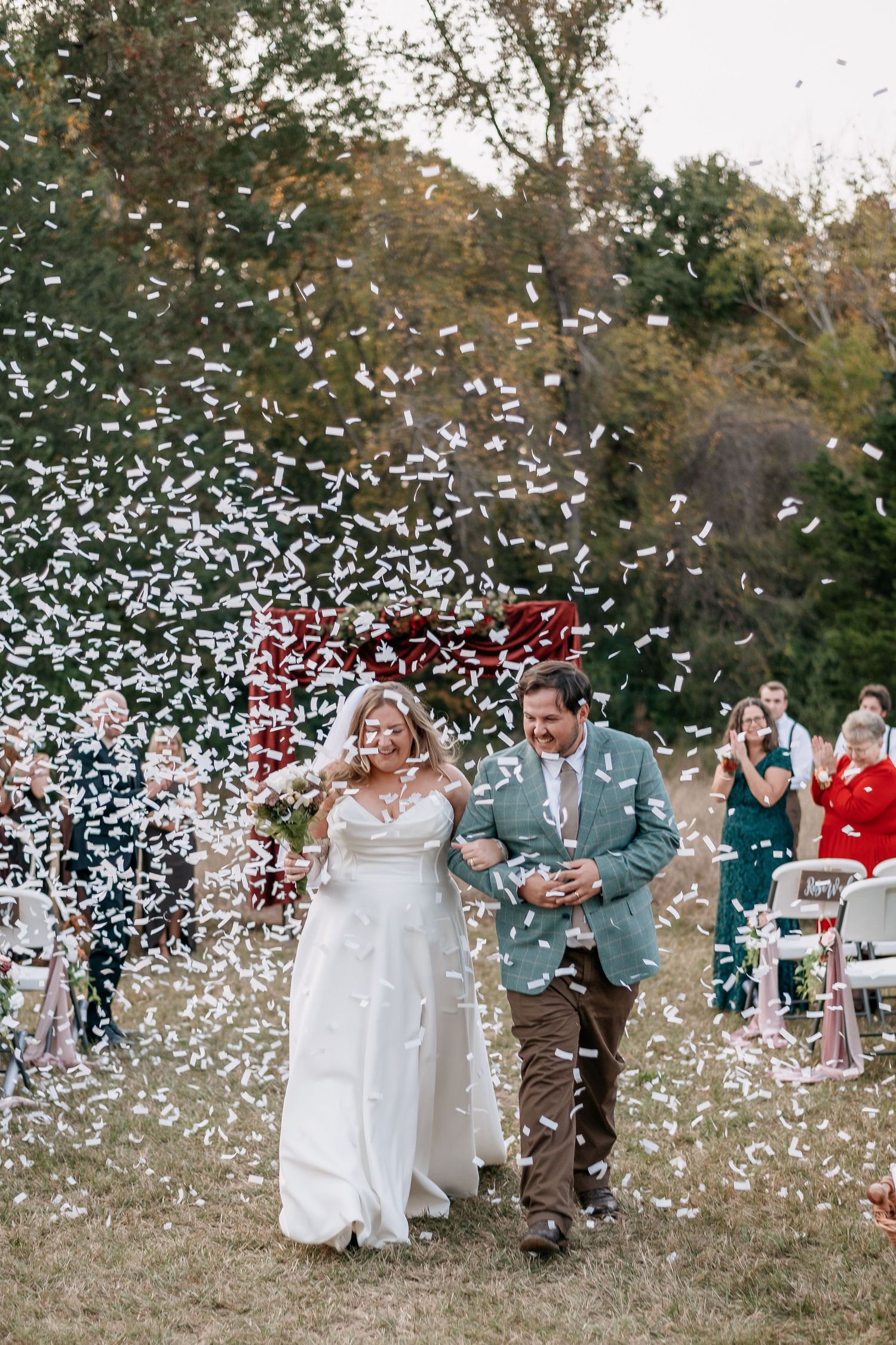 Bride and groom walk down the aisle amid white confetti shower at outdoor wedding ceremony.
