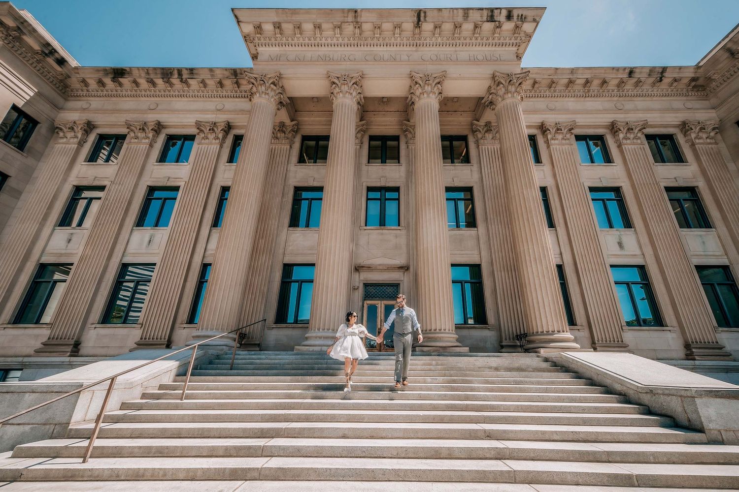 Couple holding hands on grand courthouse steps with tall columns under blue sky.