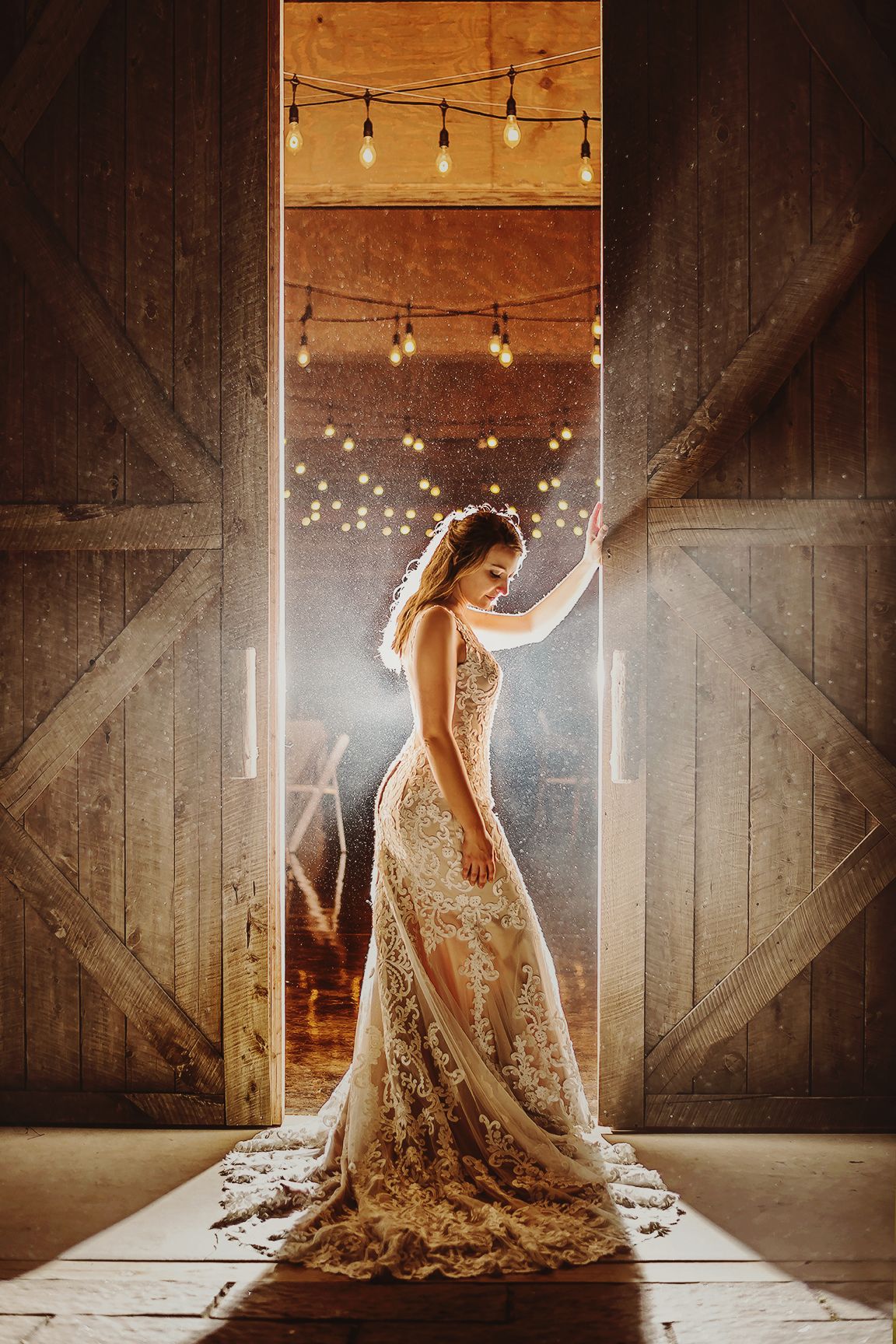 A bride in a lace wedding gown poses in a rustic barn venue with string lights and wooden walls creating a warm ambiance.