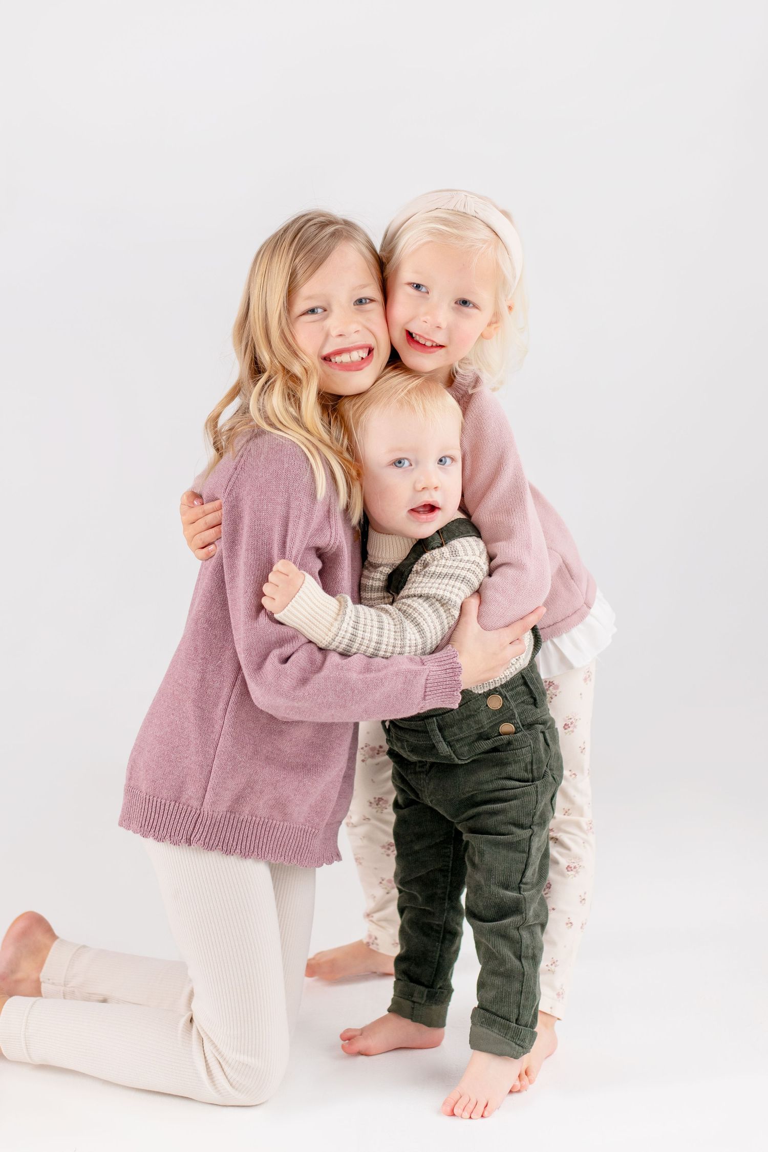 Three young children in cozy sweaters embrace and smile together against a white studio background.