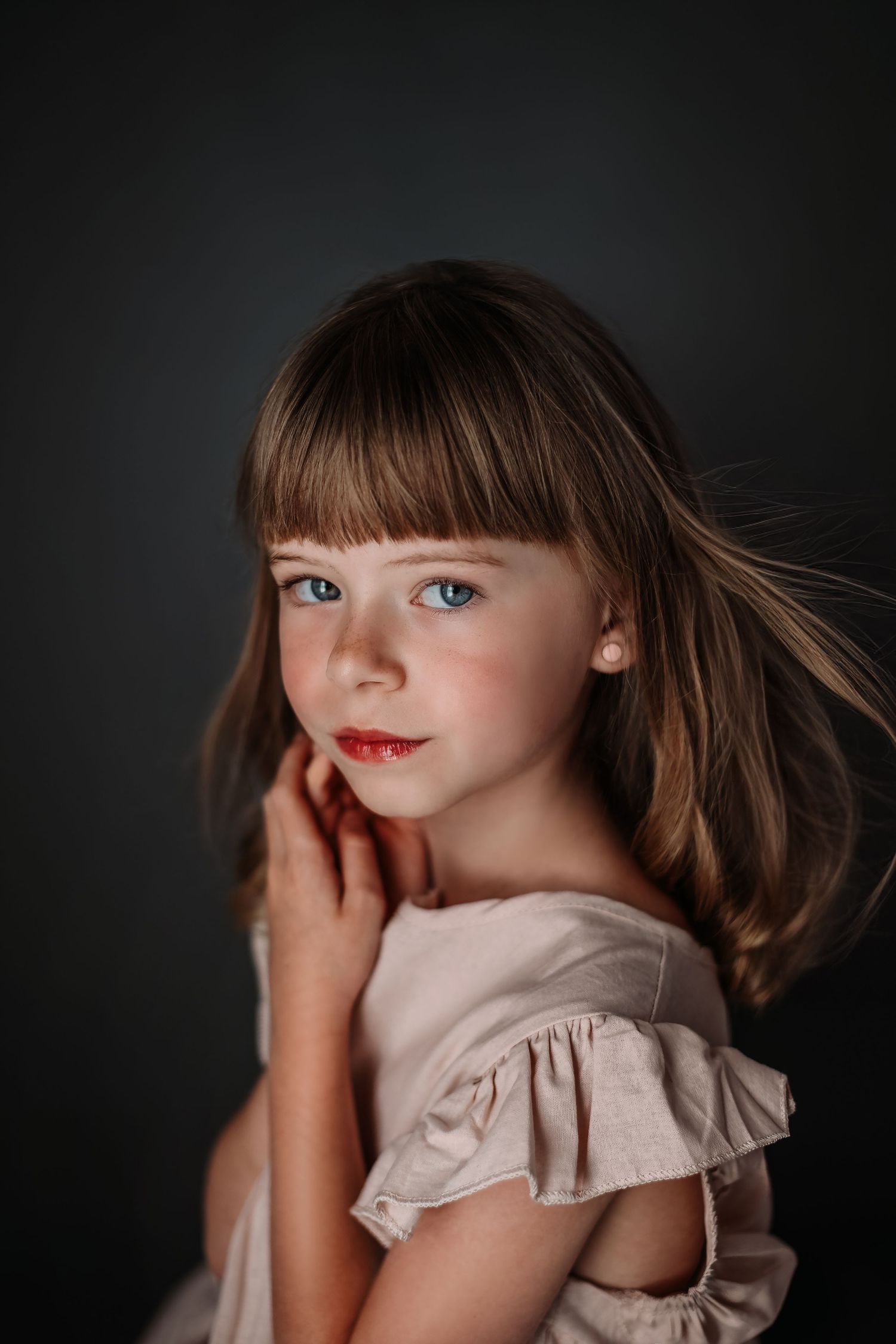 An artistic portrait showing a young person with bangs and flowing hair in a light colored dress against a dark background.