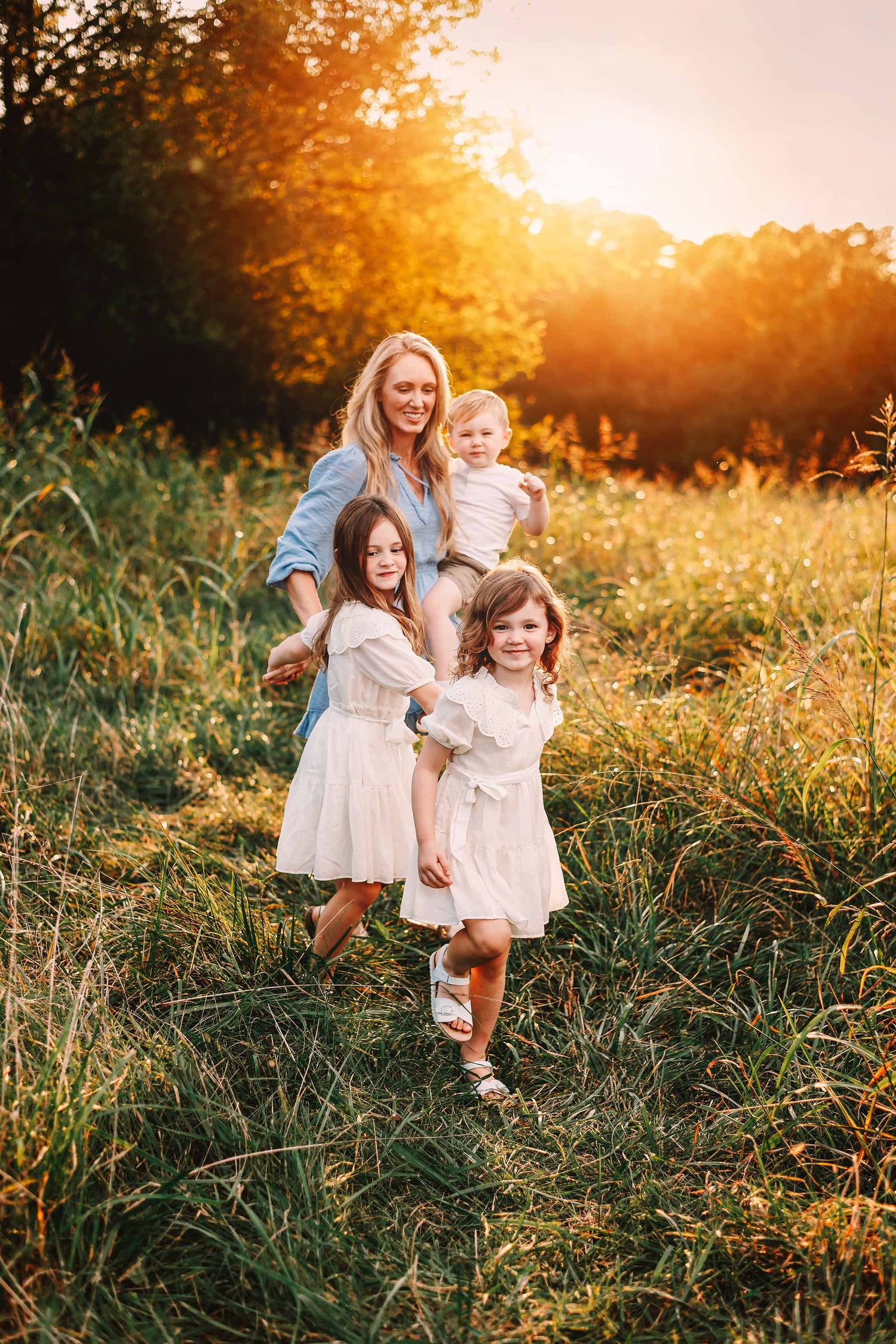 A mother in a blue dress holds a toddler while two girls in white dresses play in a sunlit meadow.