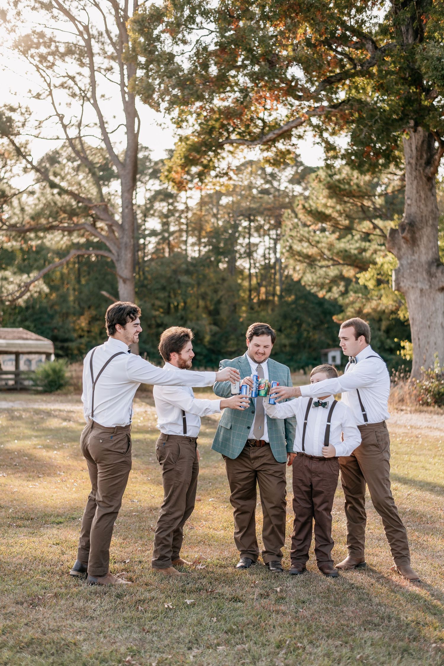 Groomsmen in brown pants and suspenders toasting with the groom outdoors under trees at a fall wedding.