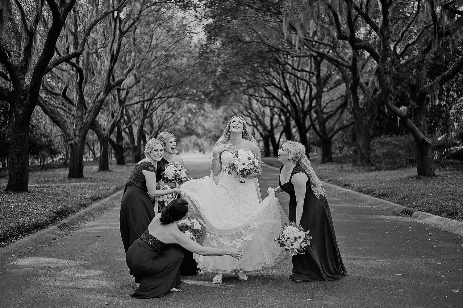 Bride and bridesmaids pose on a tree-lined path in a stunning black and white wedding photo.