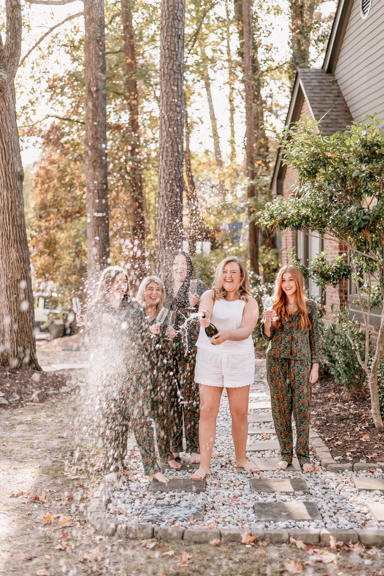 Three women celebrating outdoors, popping champagne bottle, laughing joyfully among tall trees near a house.