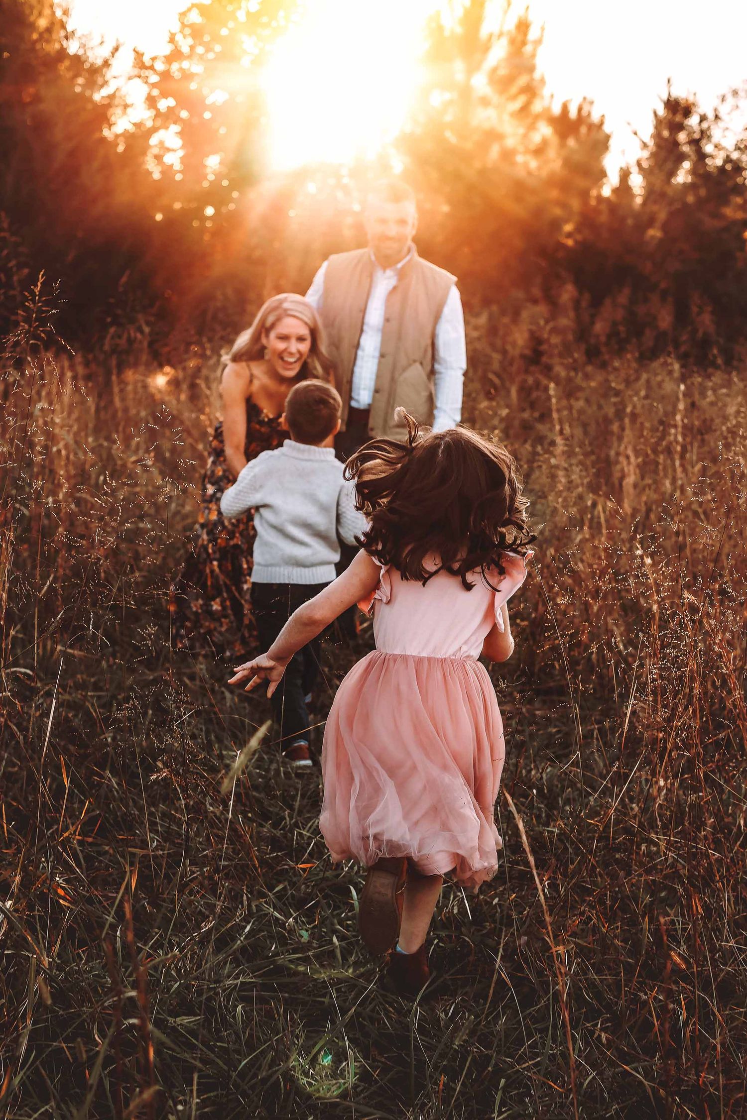 Two children run toward their parents in a golden sunset field, family embracing in warm autumn light.