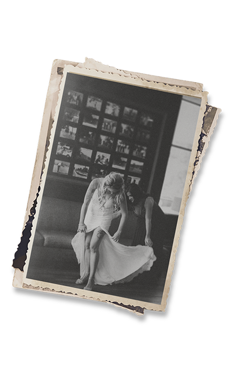 Vintage black and white photo of a woman in a white dress, wind-blown skirt, standing before a wall of framed pictures.