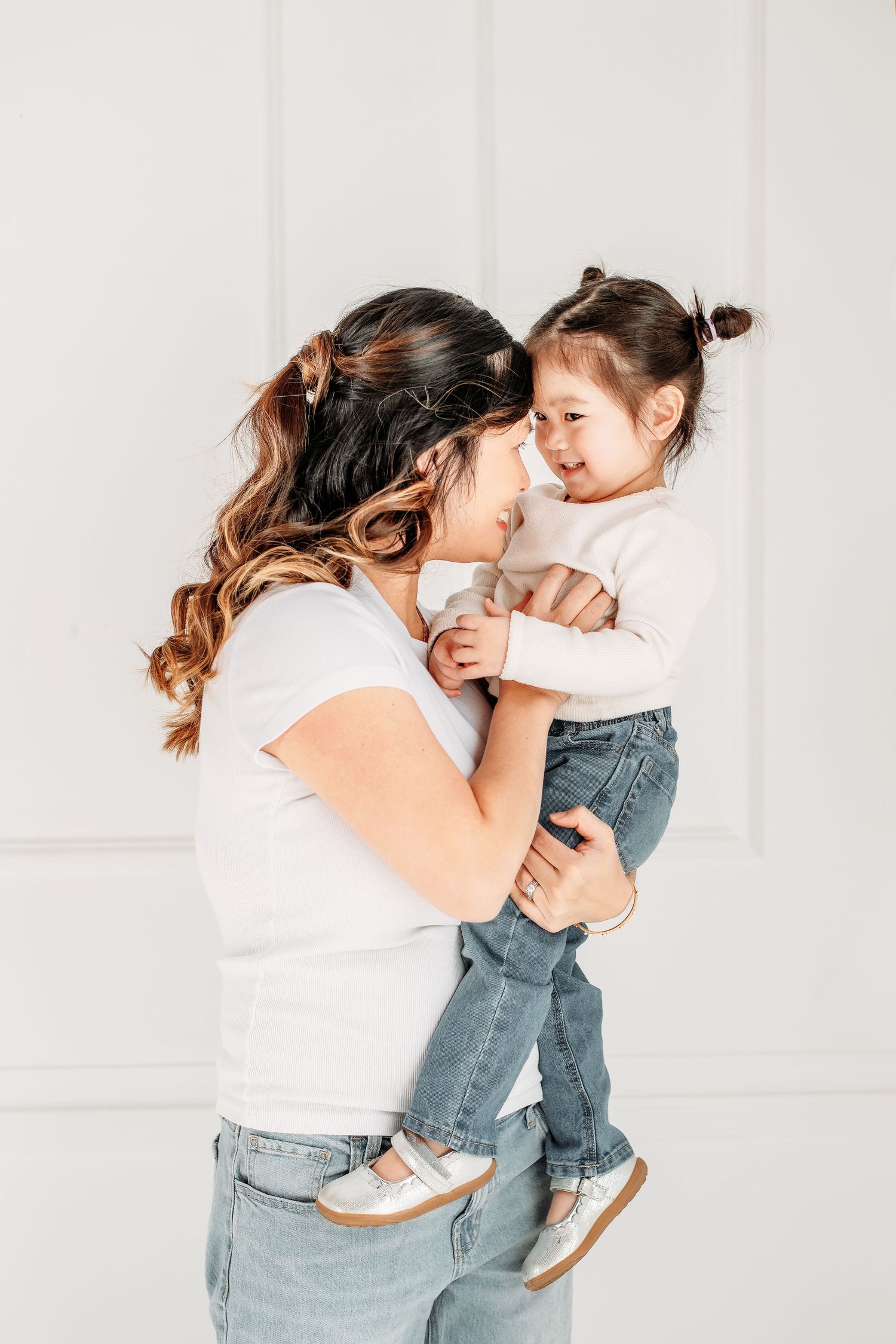 Sweet bonding moment between parent and young child both wearing white tops and denim against a light background.